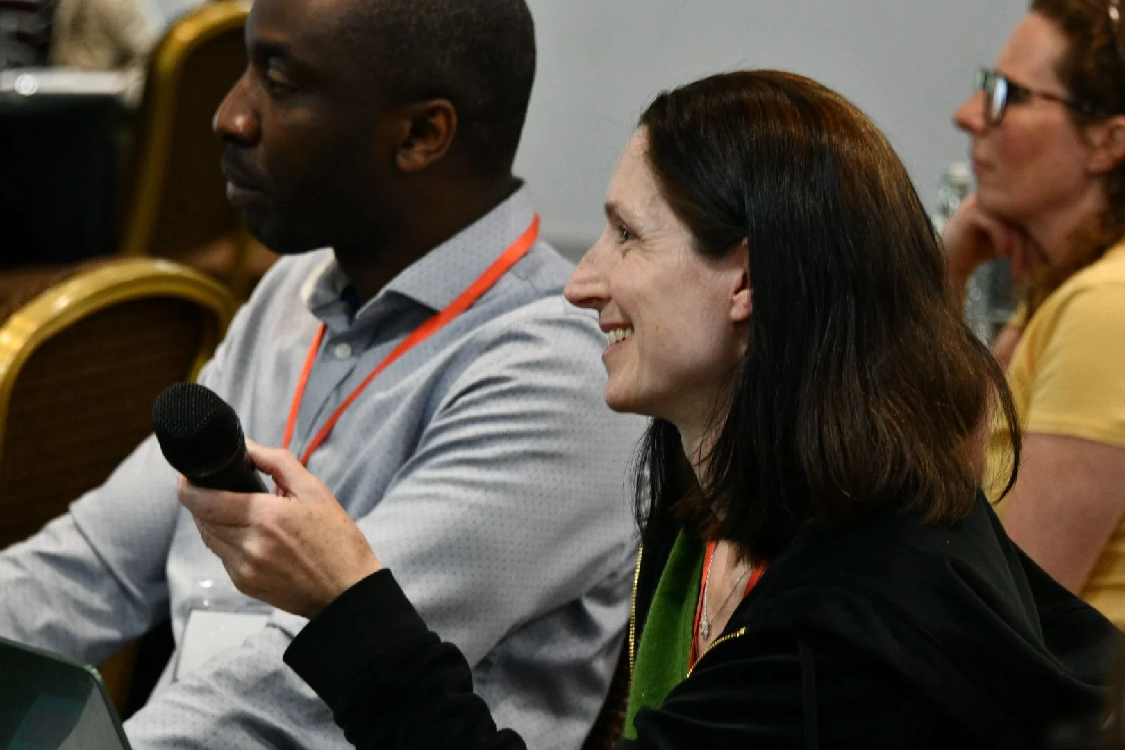Three people attending a conference or meeting. The person in the foreground is a woman with dark brown hair, smiling, wearing a black jacket and holding a microphone. The person next to her is a man with dark skin, wearing a grey shirt, also smiling, with a conference badge around his neck. The third person, partially visible in the background, is a woman with curly hair and glasses, resting her chin on her hand, wearing a yellow shirt.