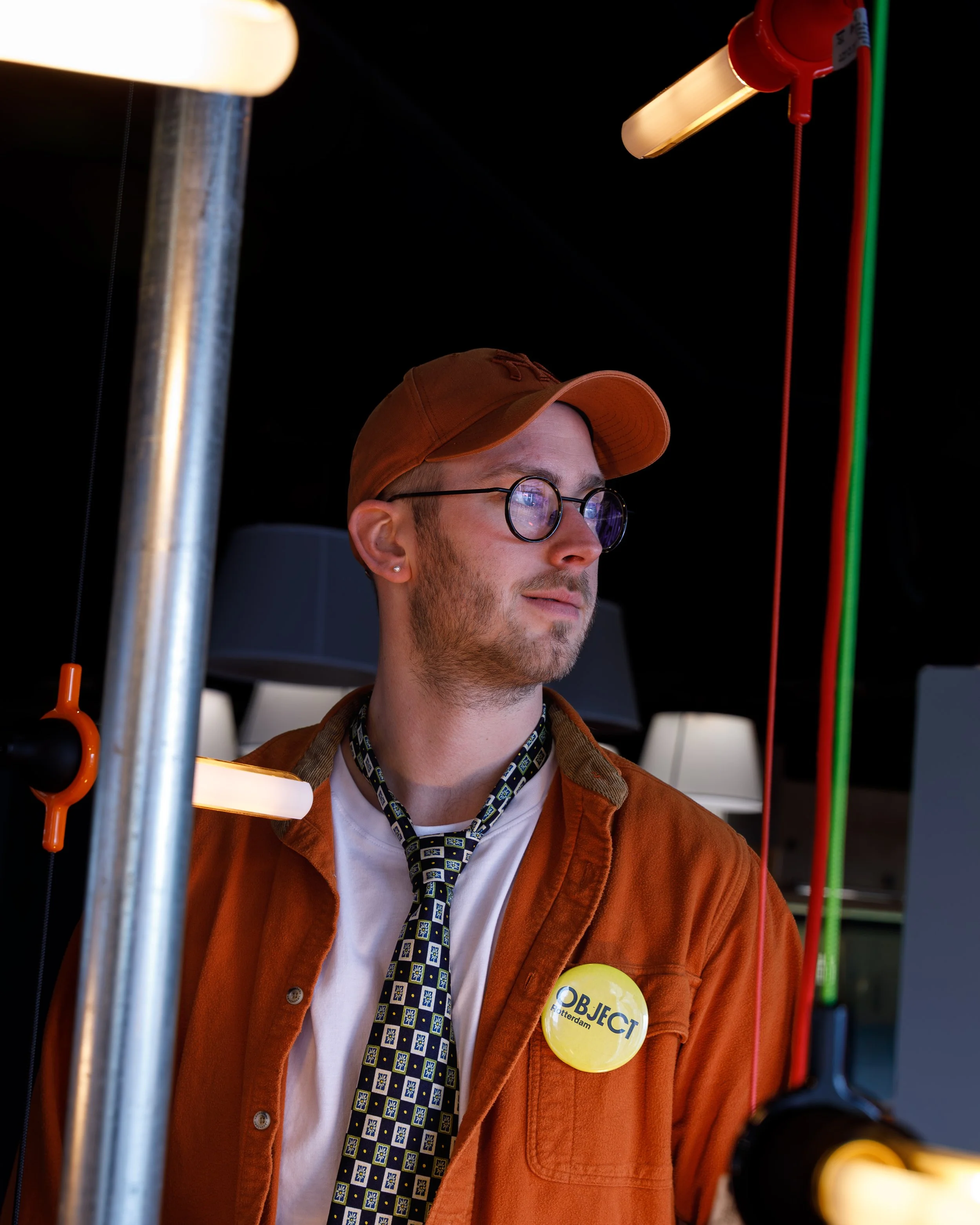 A young man with glasses, a brown cap, and earrings wearing a brown jacket with a yellow "Object" button, looking to the side among hanging lights.