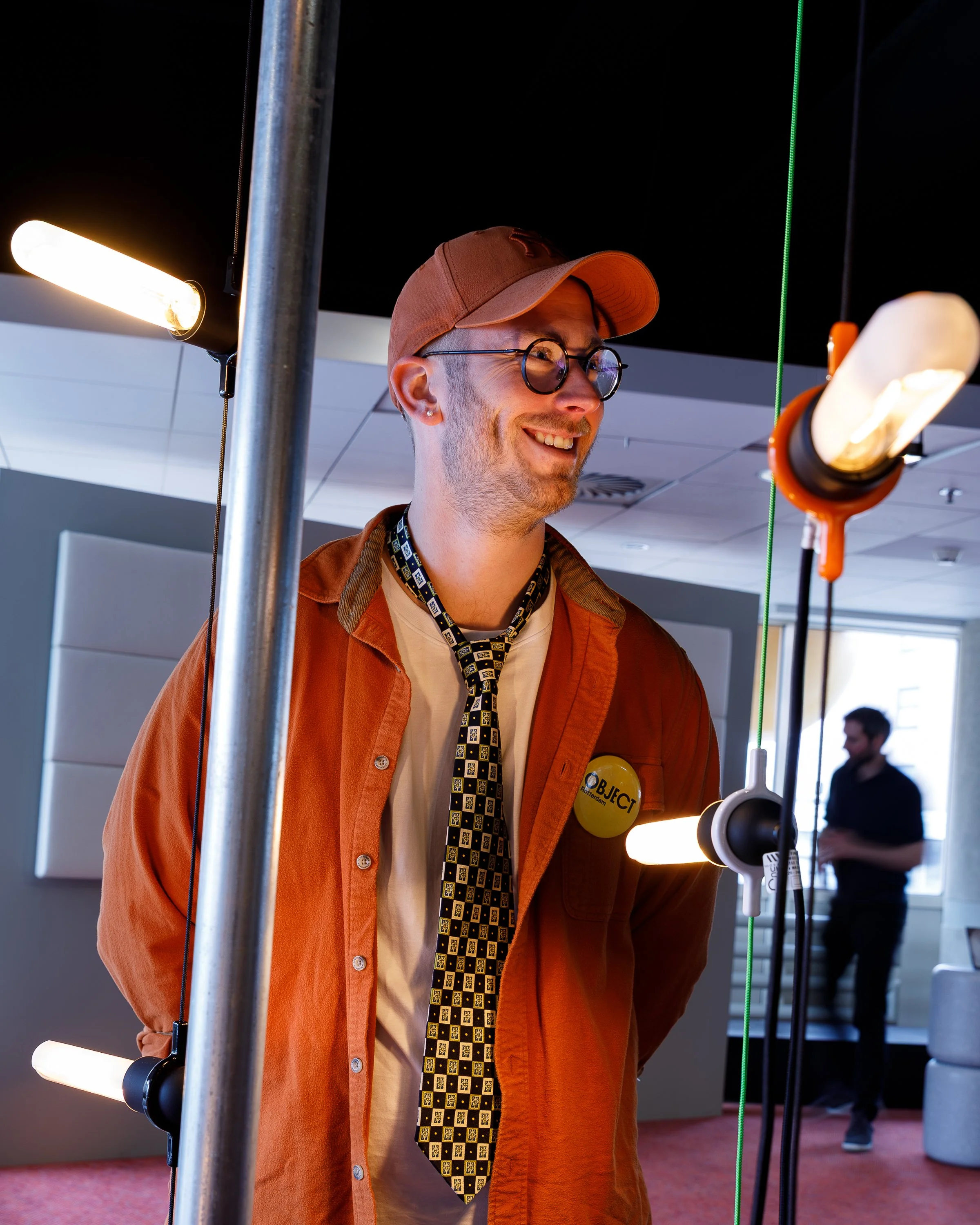 A smiling man with glasses, a brown cap, and an orange shirt, standing in a room with lighting equipment and a person in the background.