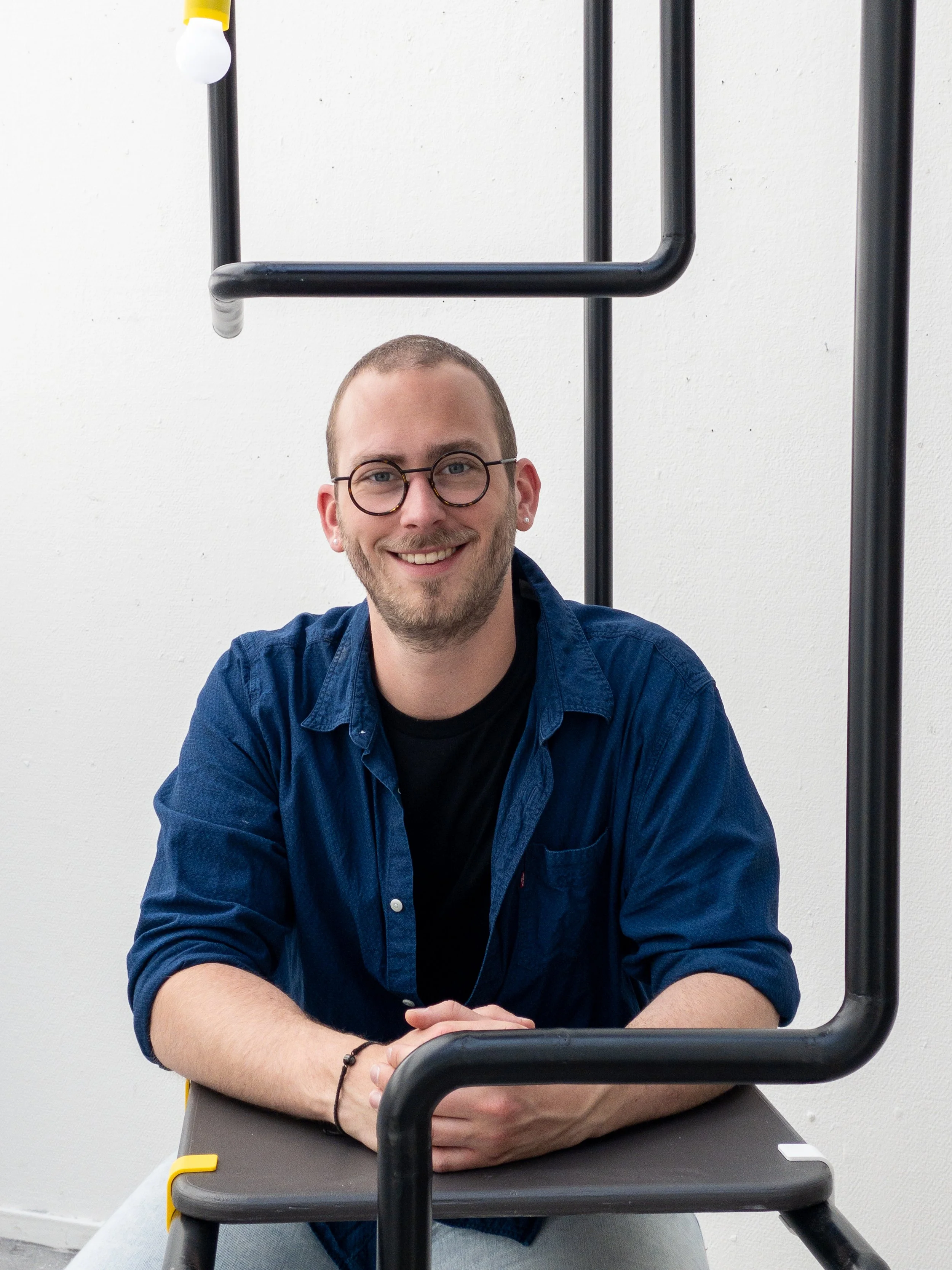 A smiling young man with glasses and a beard wearing a dark blue shirt sits at a table, with a white wall behind him and black metal pipe structure framing the image.