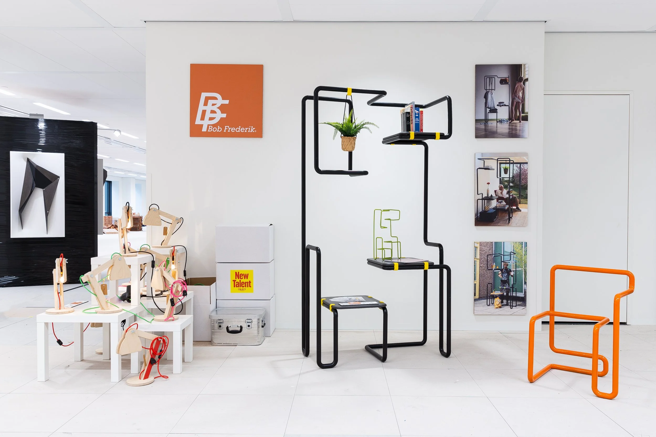 Interior display of modern furniture and decor items, including a black abstract furniture, an orange chair, and a set of small wooden desk lamps on white tables, with framed photography on the wall and a large sign that reads 'Bob Frederik'.