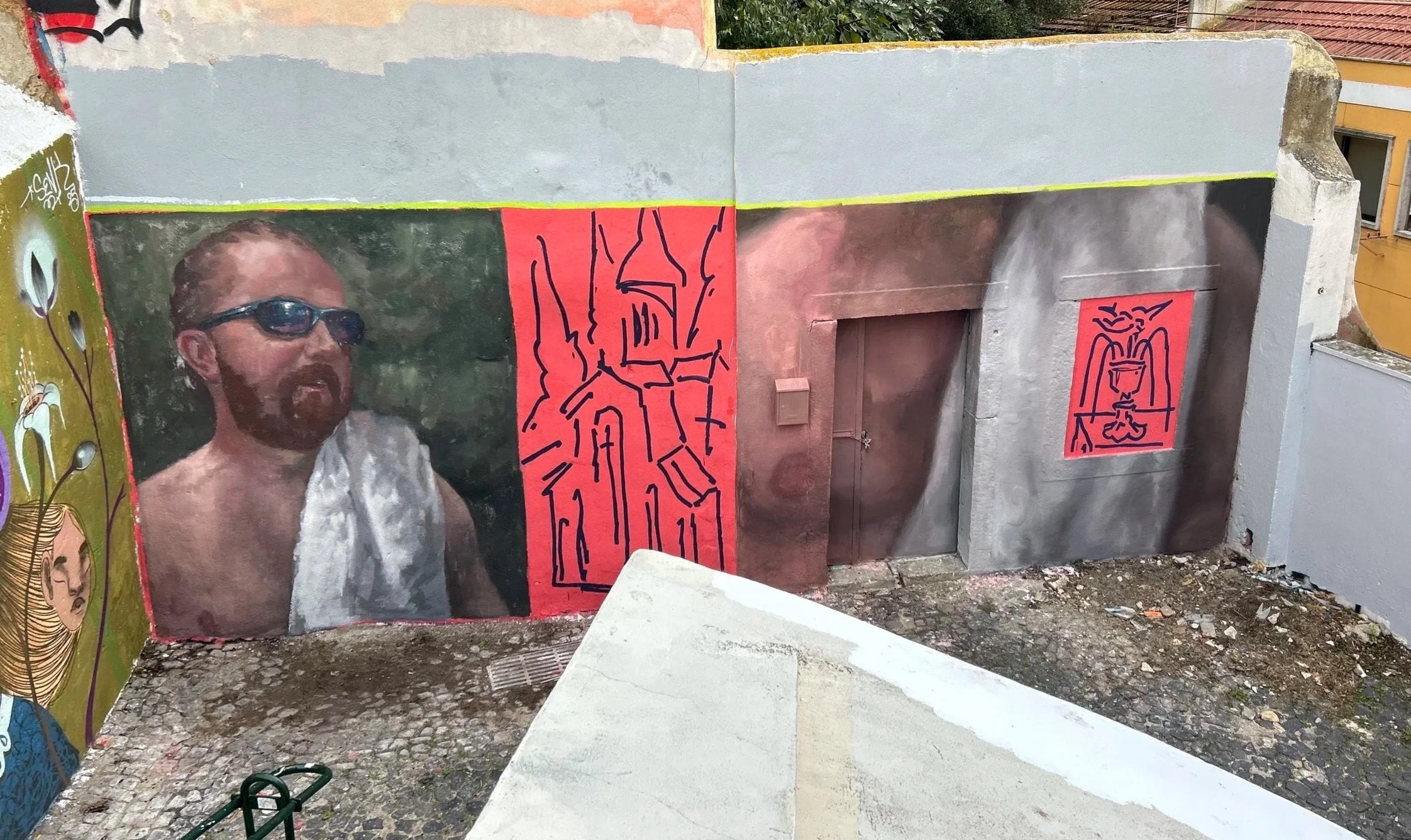 Street art mural featuring a man with sunglasses, a beard, and a towel over his shoulder on a wall, with abstract line drawings and geometric shapes in red and dark colors around him.