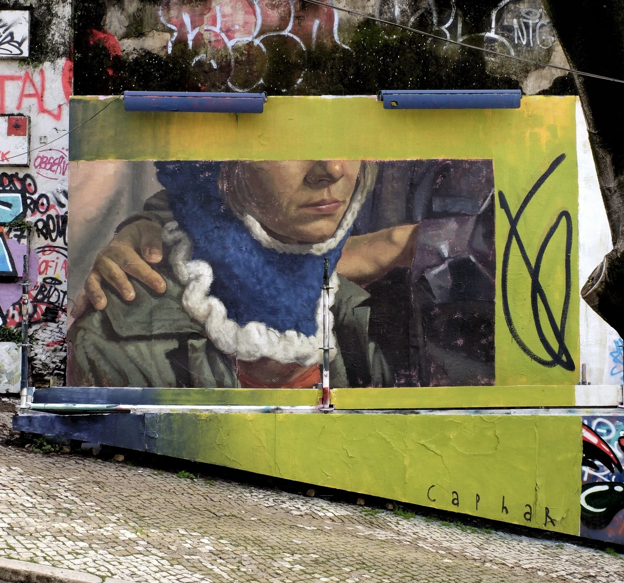 Street art mural of a woman with a fuzzy blue and white hoodie, holding a phone, painted on a wall with graffiti, signed by 'Caphar.'