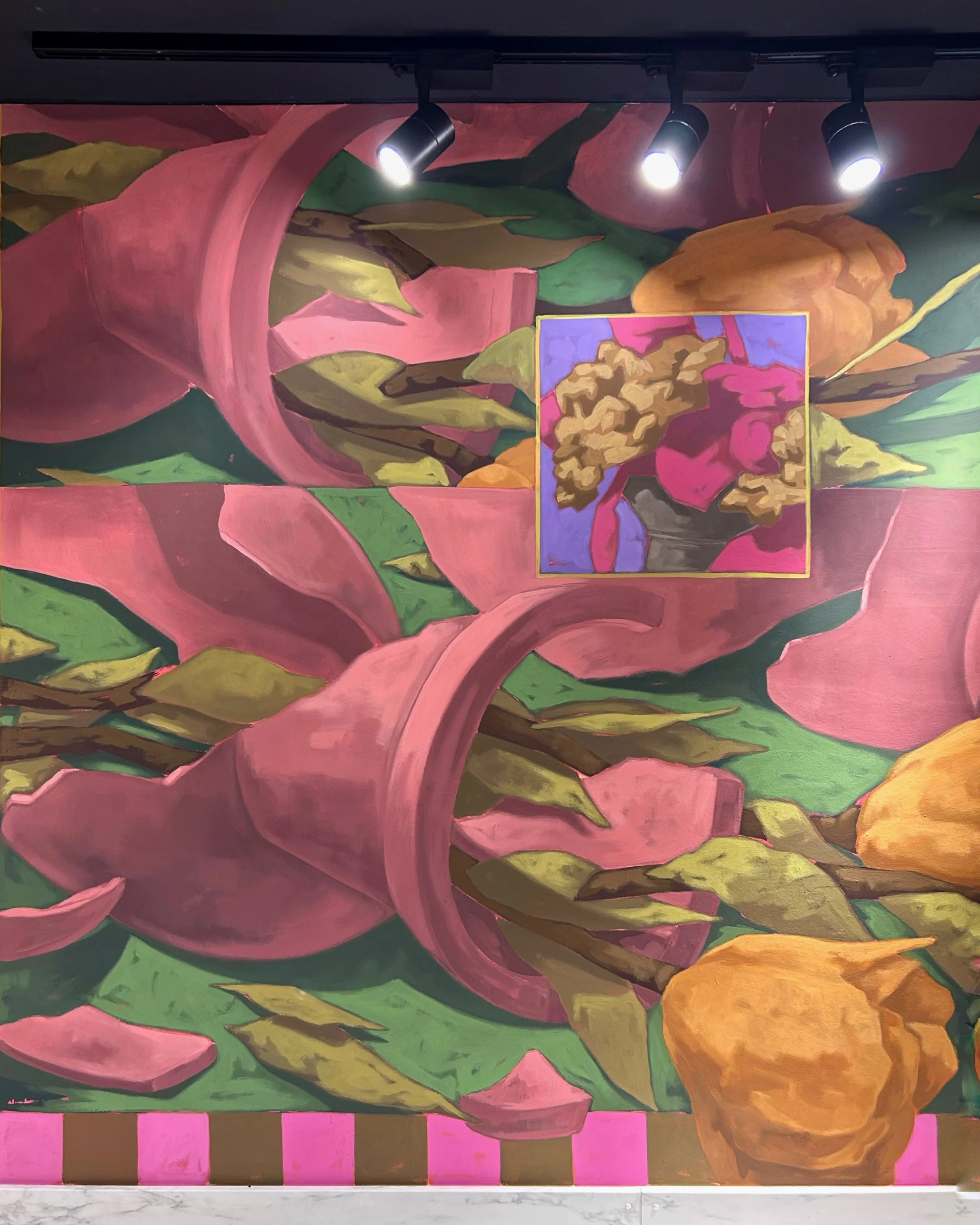 Colorful abstract floral mural with pink, green, and orange tones, featuring a framed floral painting in the center, illuminated by three overhead lights.