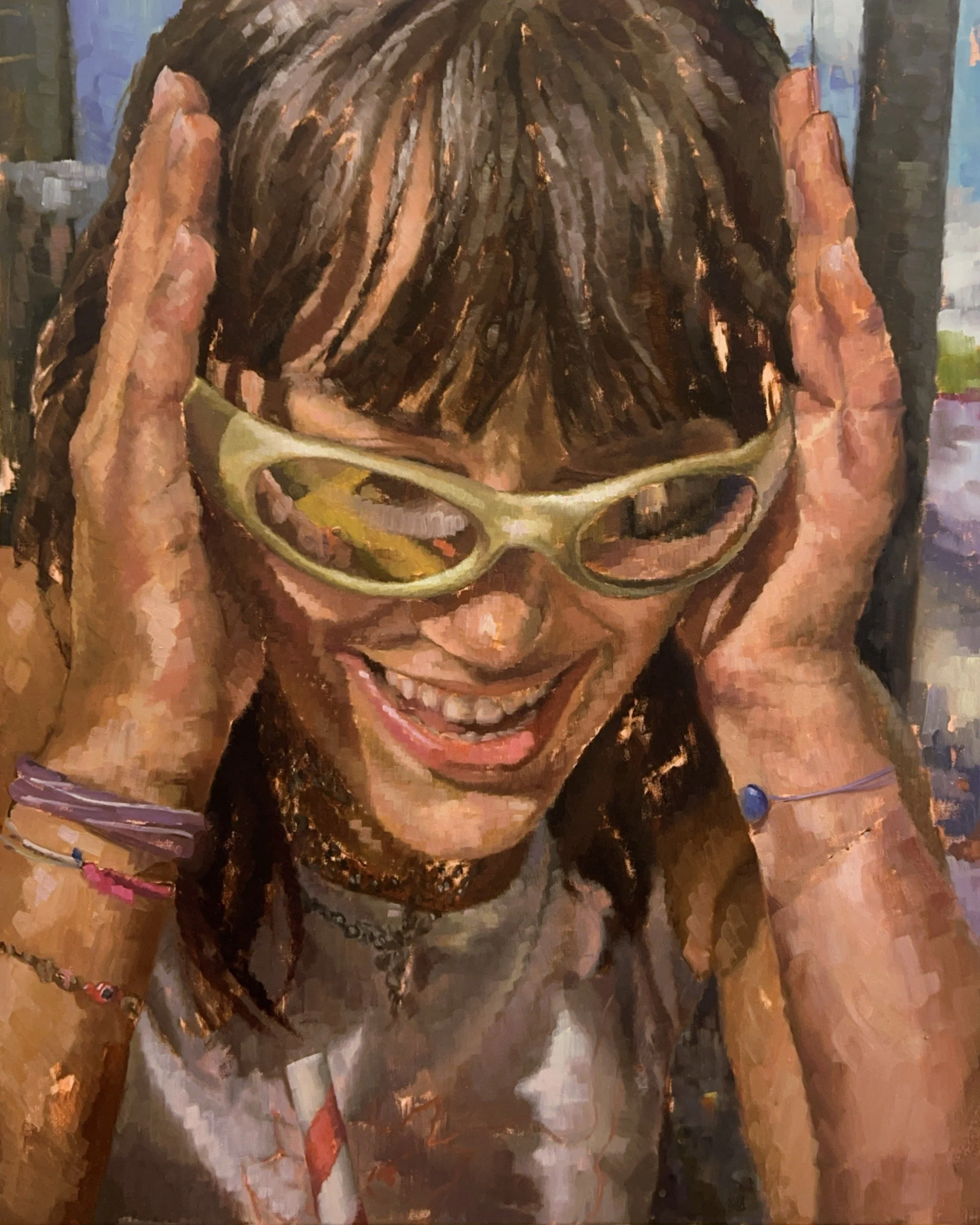 A woman with a happy expression, wearing sunglasses, with her hands on her head and smiling. She has dark brown hair with bangs and is wearing bracelets and a necklace.