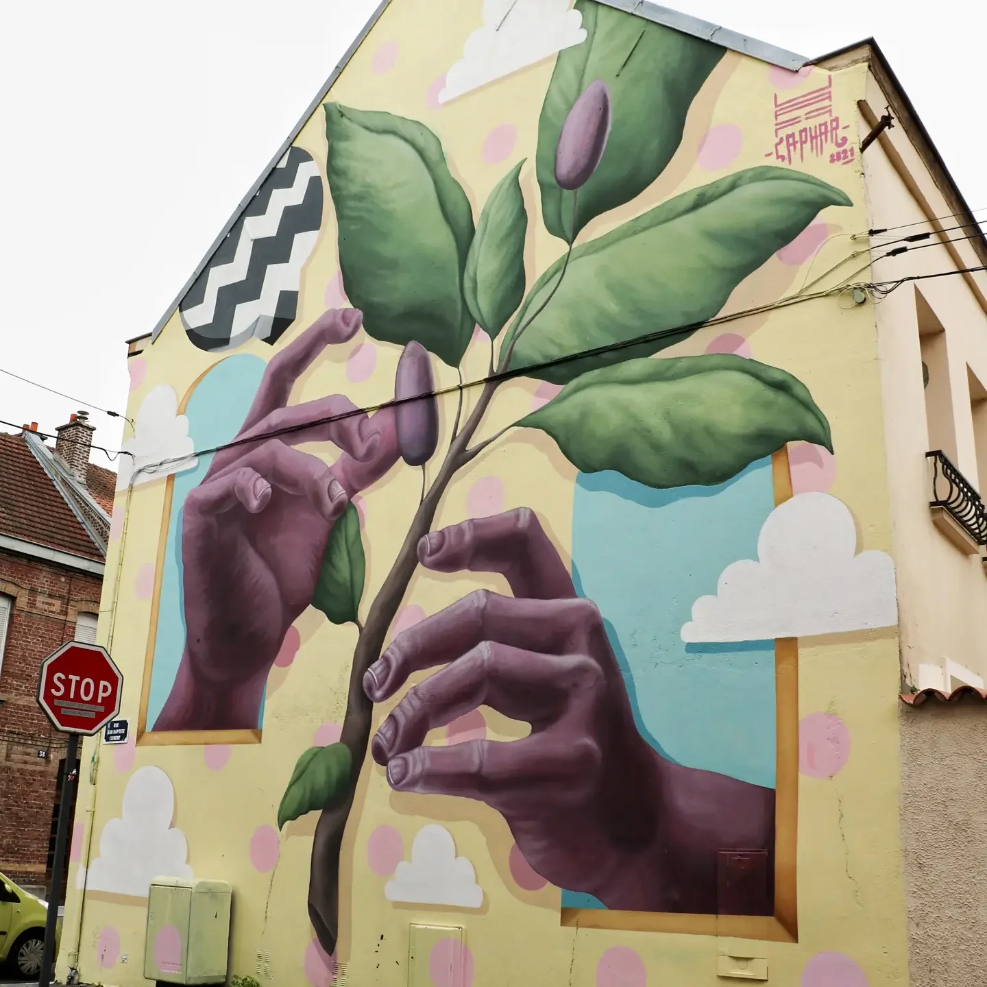 Street art mural of two realistic hands gently holding a green branch with leaves and a flower, painted on the side of a building, with clouds and pink polka dots in the background.