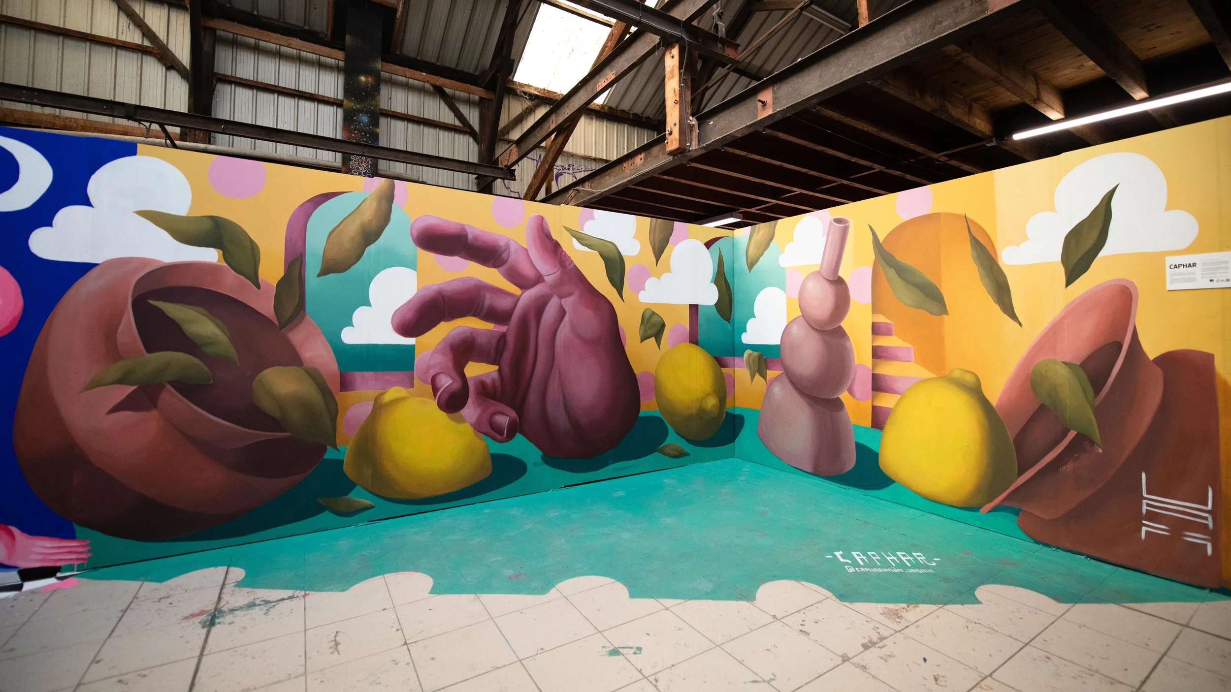 Colorful mural of a large hand reaching toward yellow pears, with a pink leaning sculpture, plant leaves, and clouds in the background.