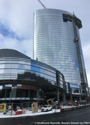A modern glass skyscraper under construction with construction equipment and vehicles at the base.