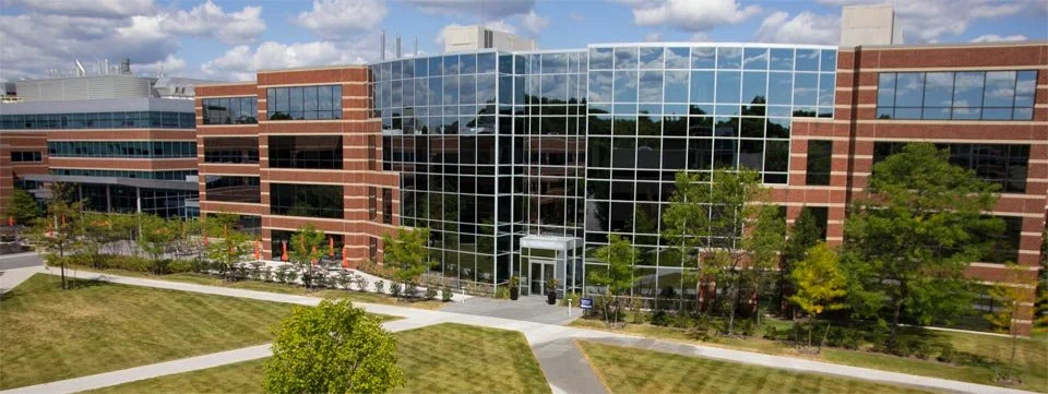 A modern office building with a glass facade, surrounded by landscaped lawns and trees, under a partly cloudy sky.