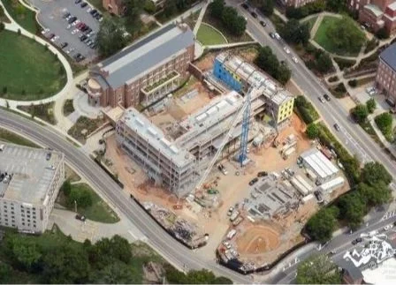 Aerial view of a construction site with a partly built multi-story building, construction cranes, and construction vehicles, surrounded by parking lots and roads.