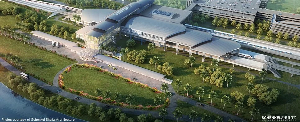 Aerial view of a modern train station with multiple platforms, surrounded by green landscaping and water nearby.