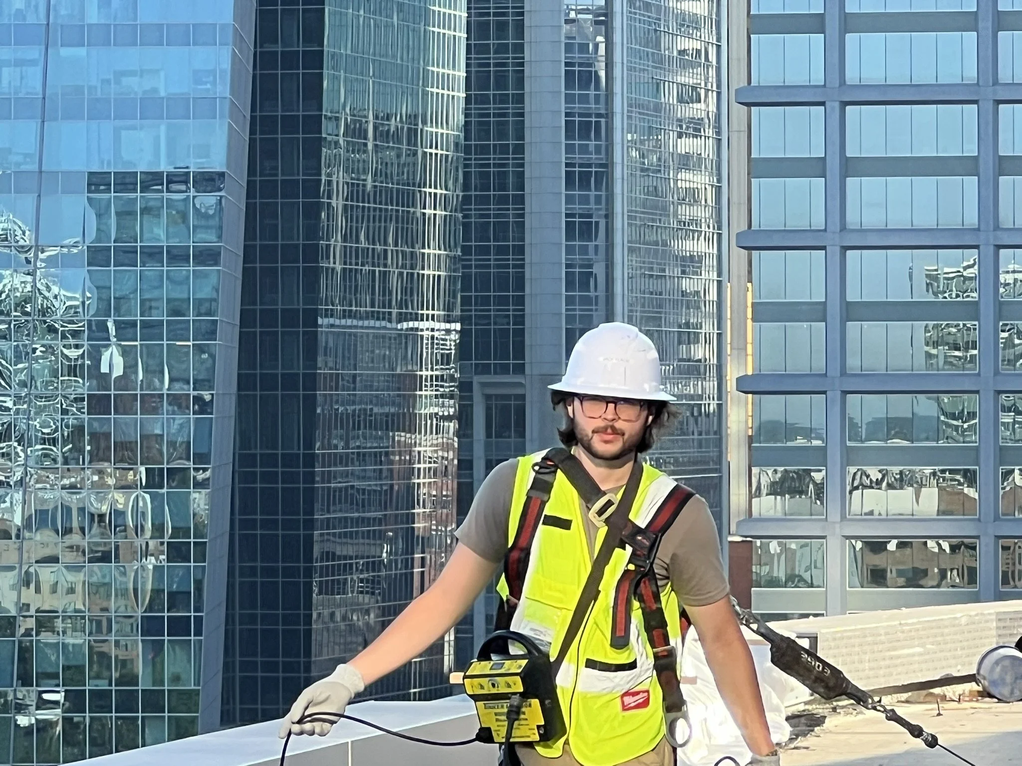 A man wearing a white hard hat, glasses, and a yellow safety vest while operating a power tool outdoors in front of modern glass buildings.