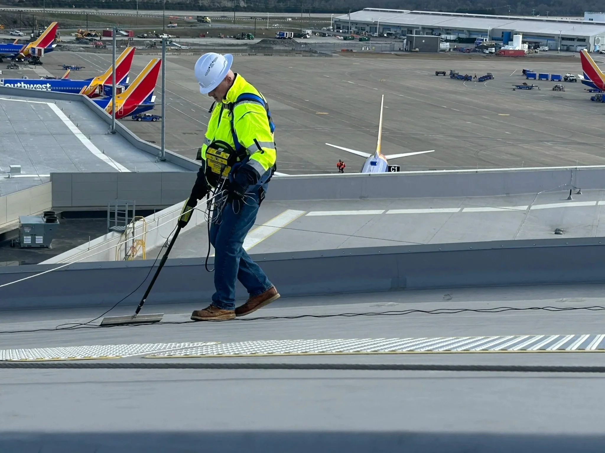 A worker wearing a white safety helmet, a yellow high-visibility jacket, and jeans is working on the rooftop of an airport terminal, handling cables or equipment near the edge of the roof. The airport tarmac with aircraft, vehicles, and airport buildings is visible in the background.