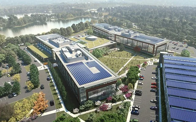 Aerial view of a modern campus with multiple buildings covered in solar panels, surrounding parking lots, green lawns, and a nearby river and forest.