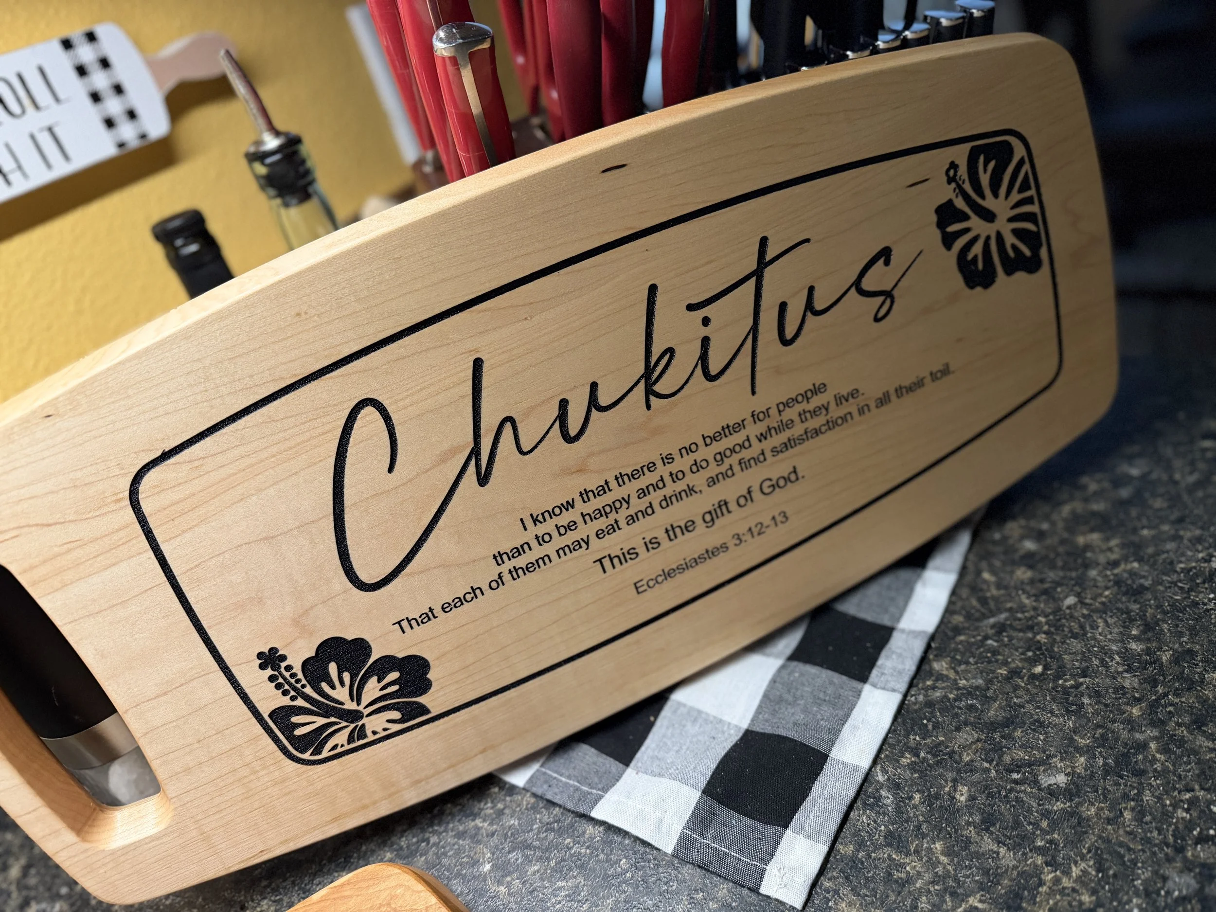 A wooden tray with the word 'Chukkutus' printed on it, along with a Bible verse from Ecclesiastes 3:12-13. The tray has decorative floral designs on each corner and is placed on a checkered cloth on a kitchen counter. In the background, there are bottles and utensils.
