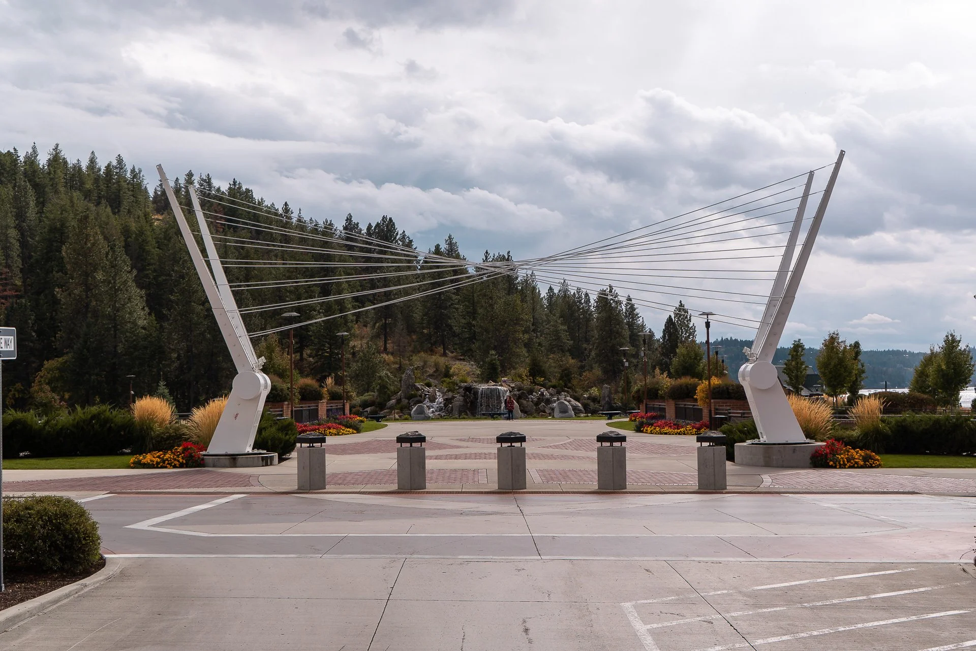 Public park with large modern street art sculpture resembling a giant pair of headphones, surrounded by landscaped flower beds, trees, and a waterfall in the background under cloudy sky.