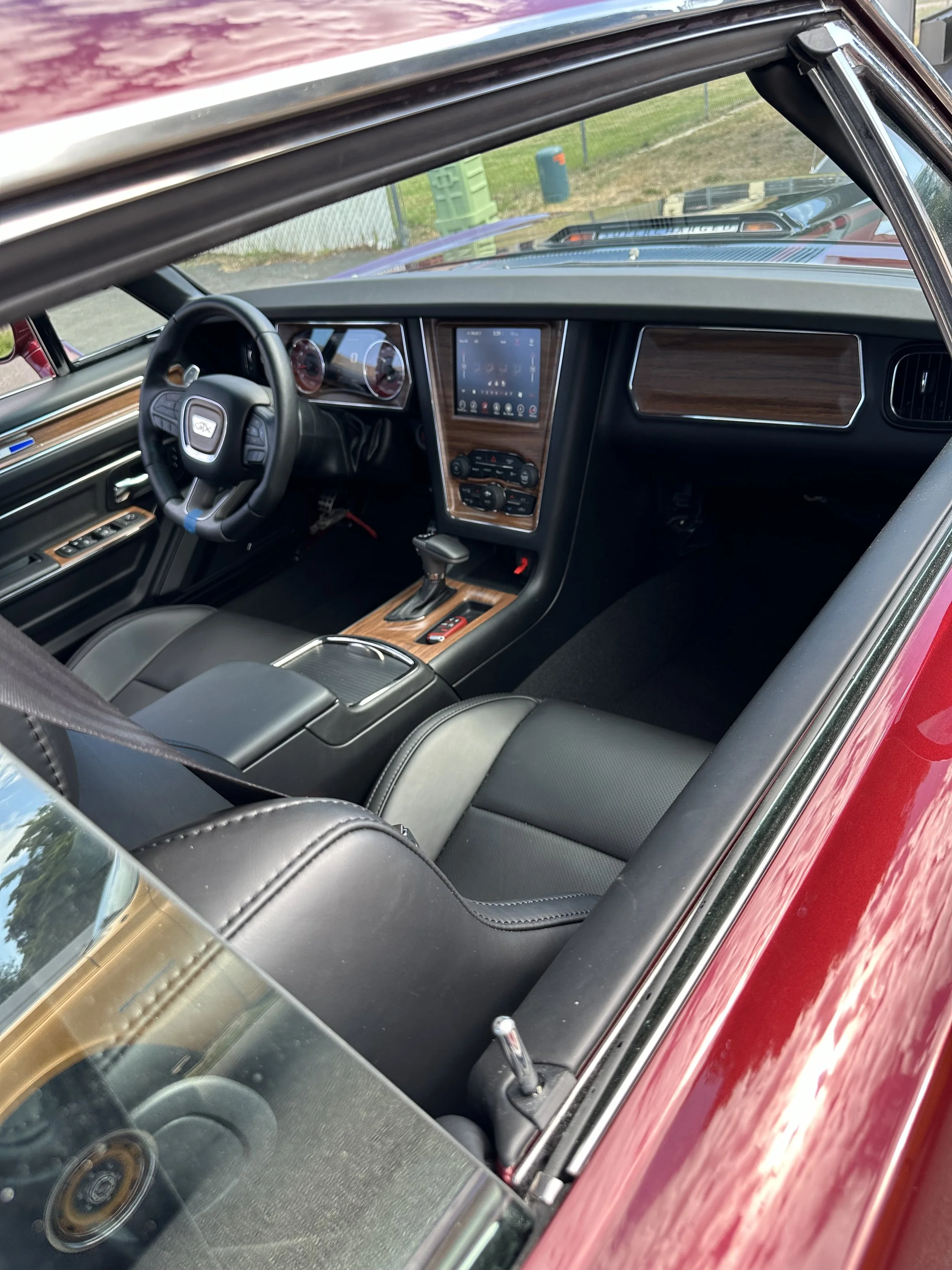 Interior view of a classic red car showing black leather seats, a dashboard with a touchscreen display, wood paneling, and a steering wheel with multiple controls.