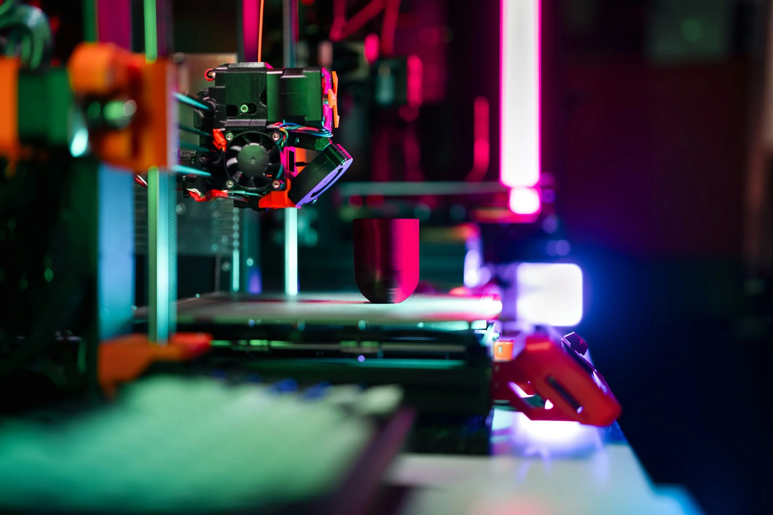 Close-up of a 3D printer in action, printing a dark-colored object with vibrant neon pink, purple, and blue lights in the background.