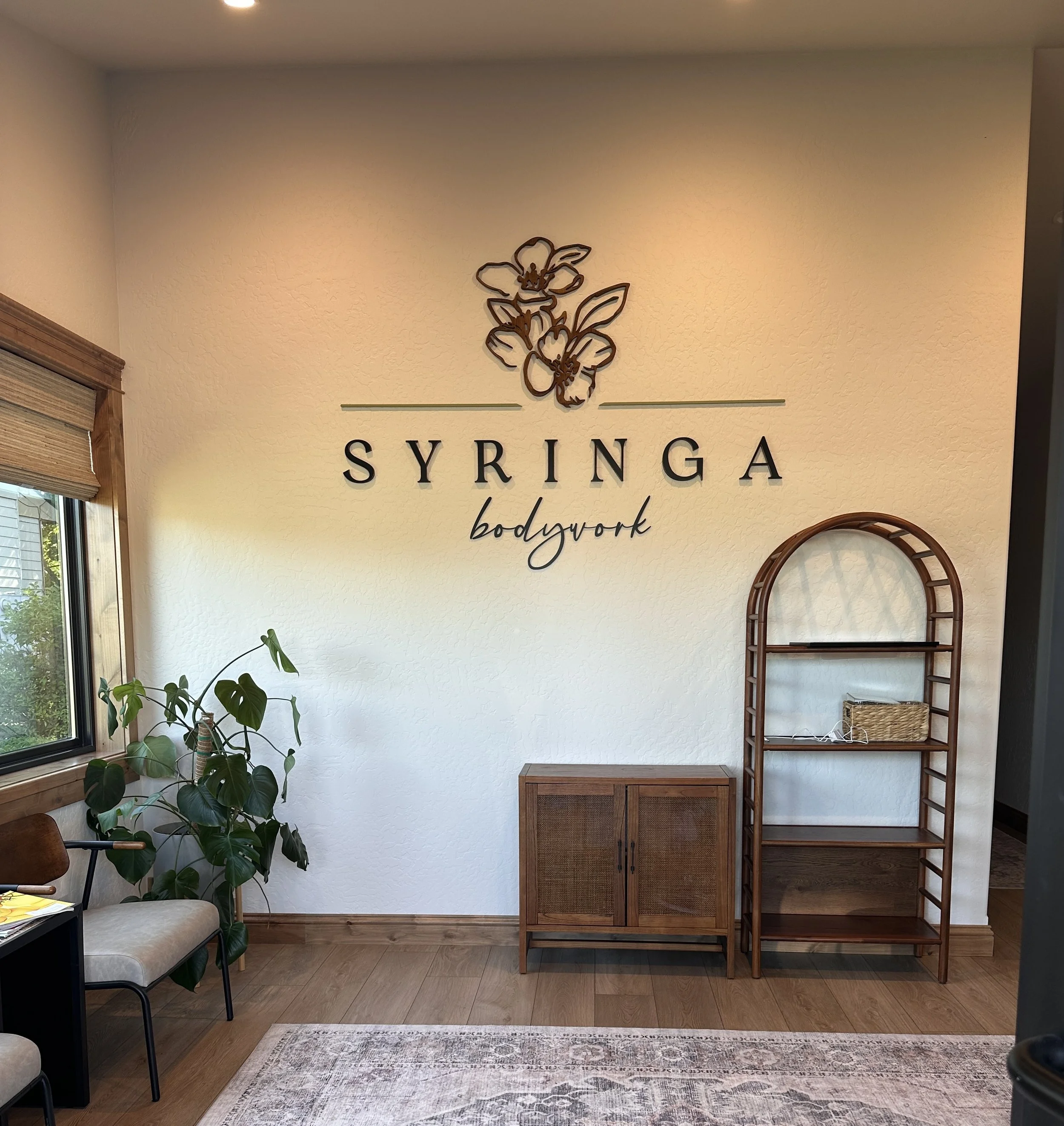 Interior of a wellness or fitness studio with a wall sign reading 'SYRINGA bodywork', featuring a flower logo above. There is a large plant near a window, a small cabinet, and a tall wooden shelf on the right.