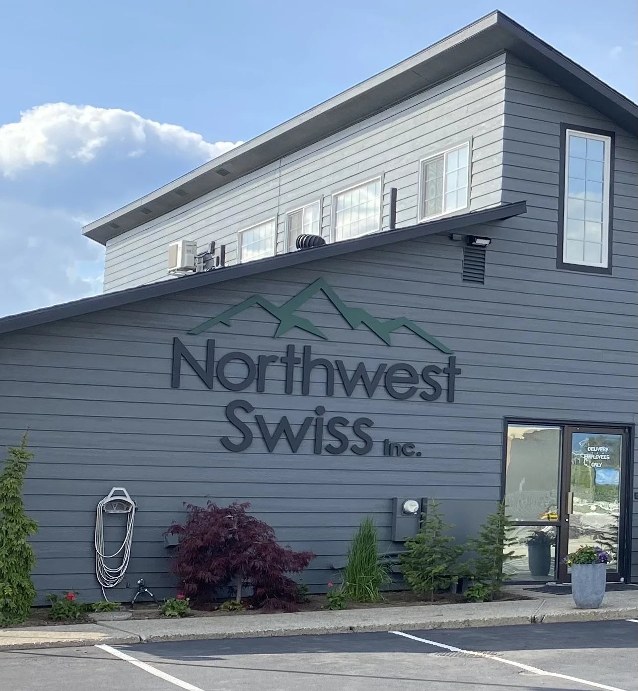 Exterior view of a gray building with a sign that reads "Northwest Swiss Inc." and features a mountain logo. The building has several windows, an air conditioning unit, and a landscaped area with small trees, plants, and a parking lot.