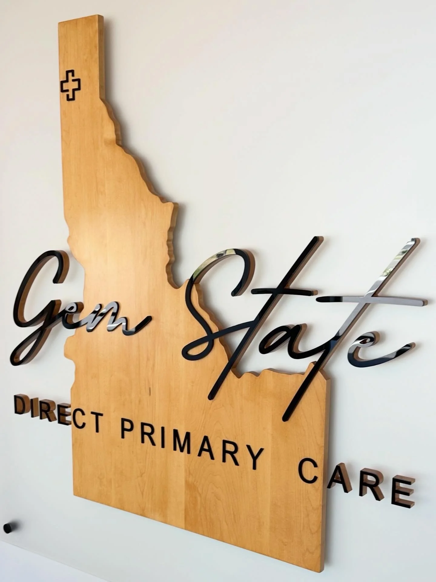 Wooden sign shaped like the state of California with black text reading "Gen San" and partially visible words "DIREC T PRIMARY CARE" hanging on a white wall.