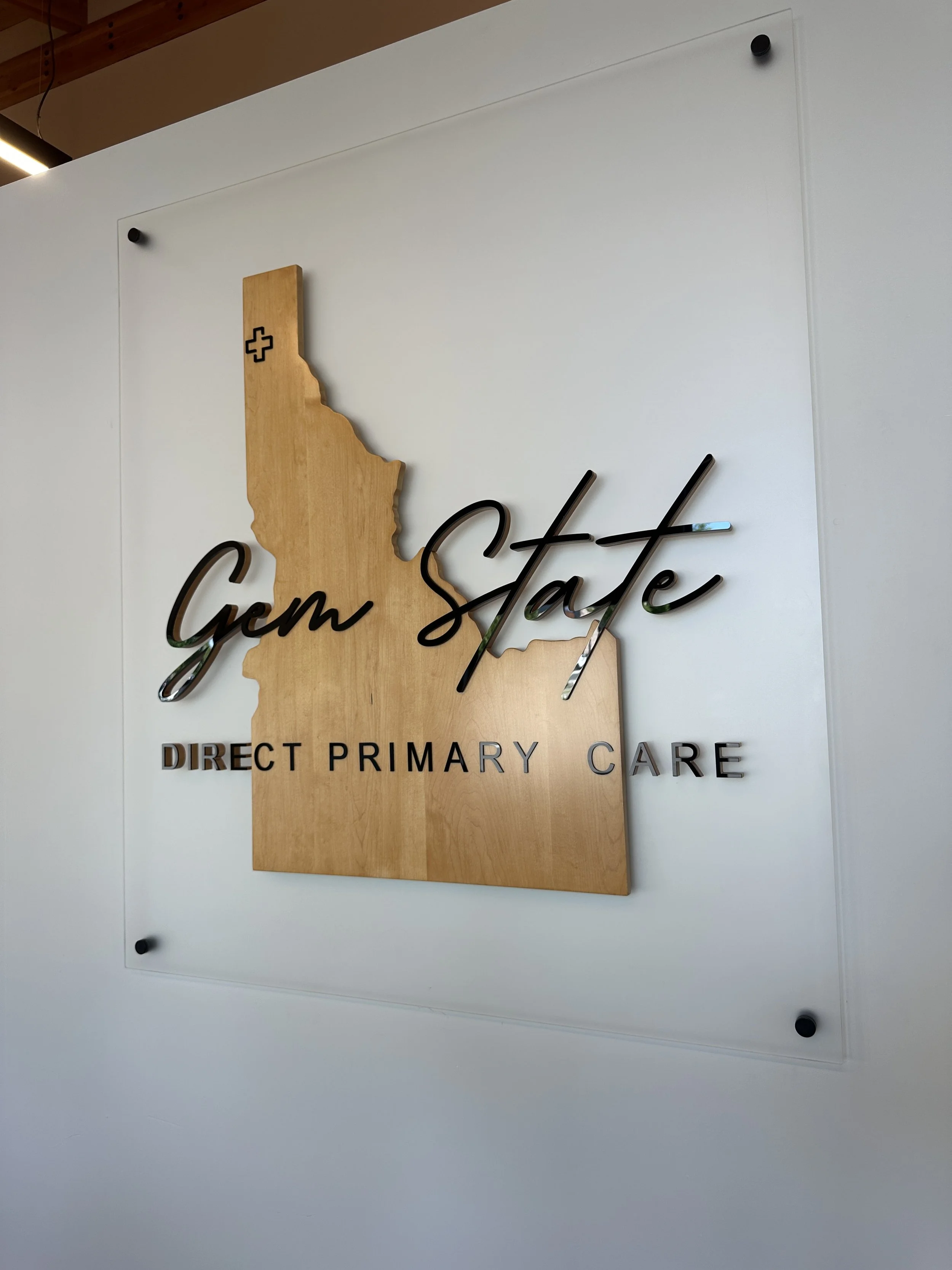 Sign for Gem State Primary Care featuring a wooden outline of Idaho with a plus symbol, and black cursive text reading 'Gem State' alongside black uppercase text reading 'Direct Primary Care'.