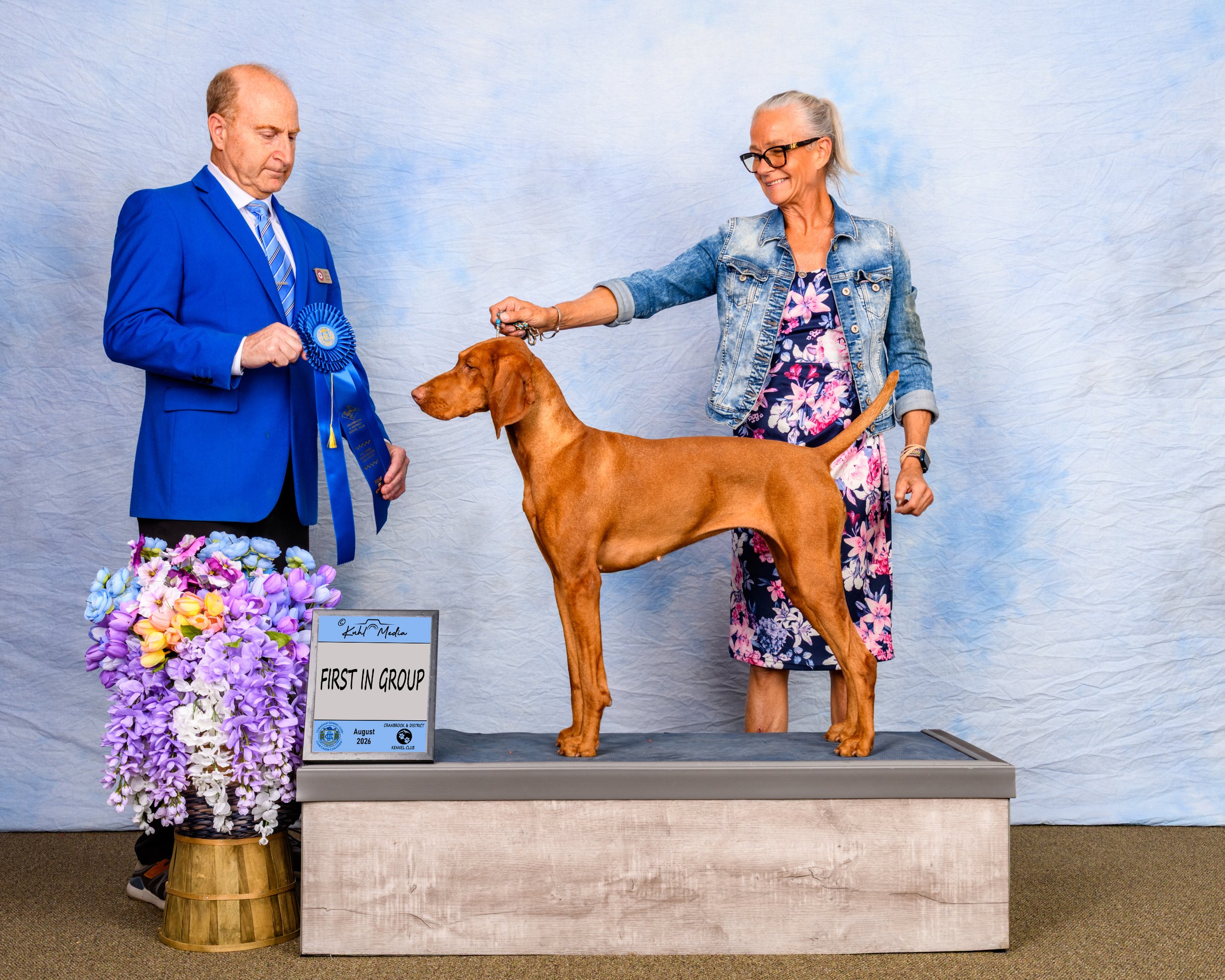 A woman and a man with a dog at a dog show. The woman is holding the dog's leash, and the man is wearing a blue ribbons. There is a flower arrangement and a sign that reads 'First in Group, August 2026'.
