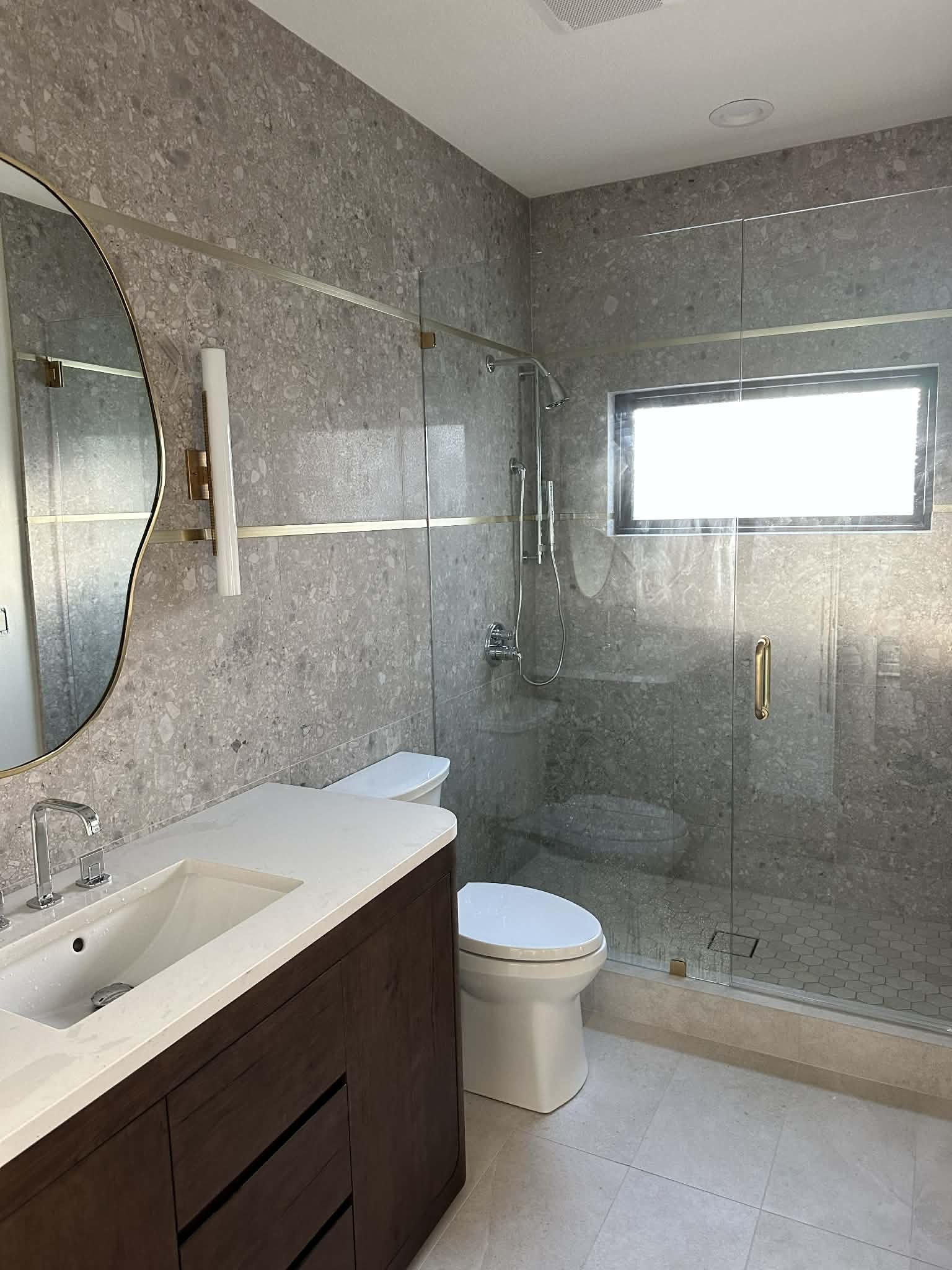 Modern bathroom with gray marble walls and glass shower enclosure, white toilet, wooden vanity with a white sink, and a mirror. There is a window with natural light.