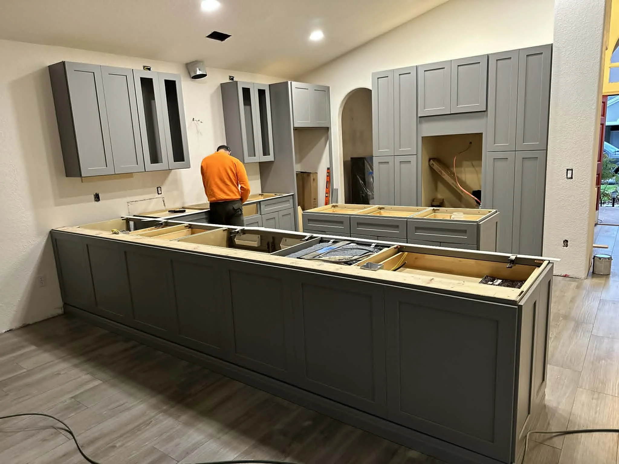 Kitchen with gray cabinets being installed, a worker in an orange shirt working at the countertop area, and construction materials on the kitchen island.