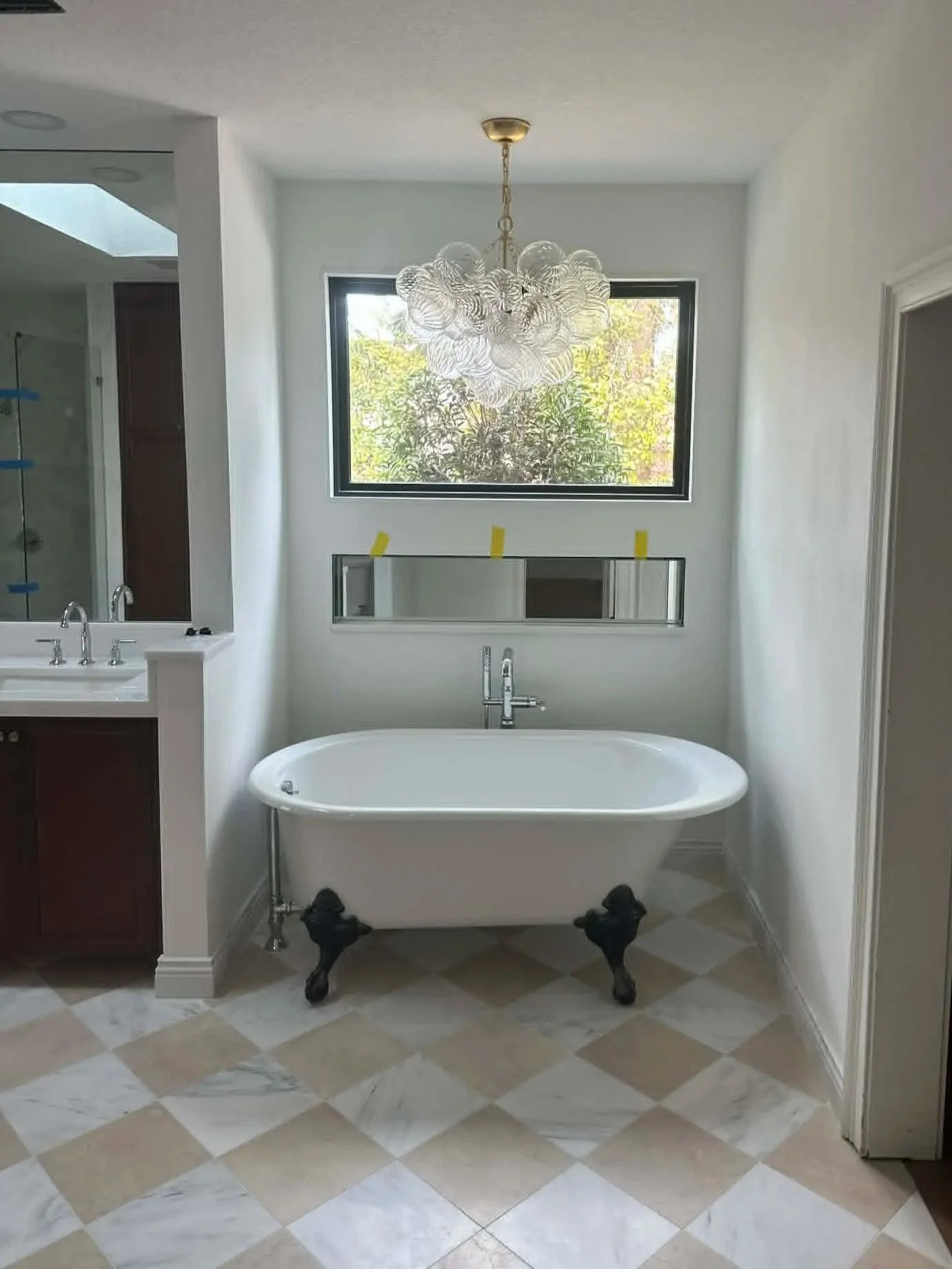 A bathroom with a freestanding clawfoot bathtub beneath a large window, with a modern chandelier hanging from the ceiling, and tiled flooring.