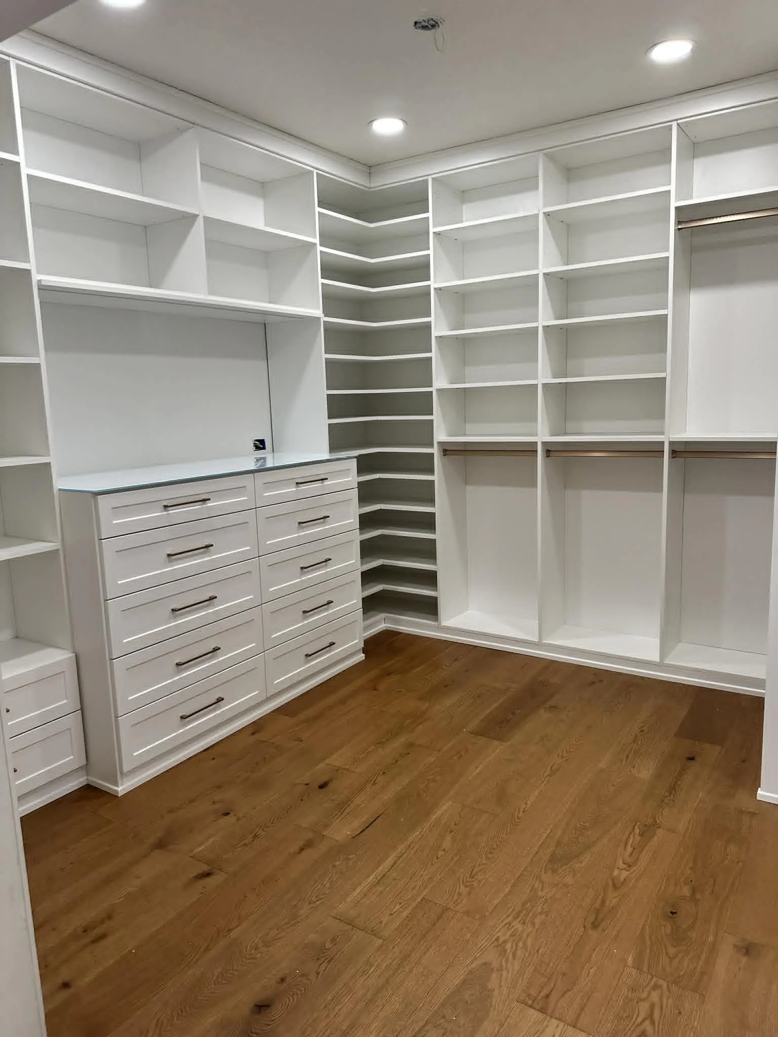 Empty white walk-in closet with built-in shelves, drawers, and wooden floors.