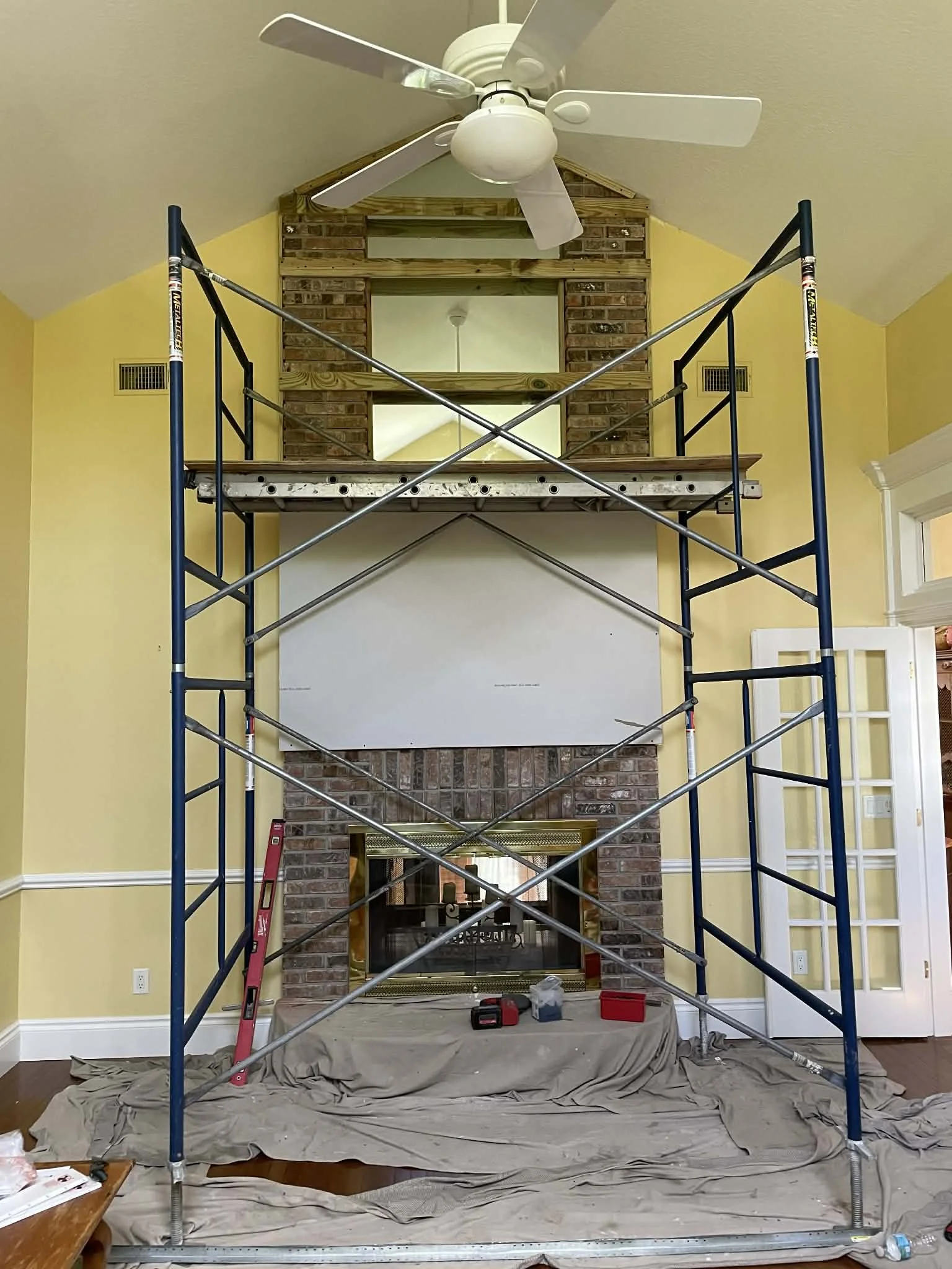 The image shows a partly constructed brick fireplace with a mirror above it, inside a room with yellow walls. A metal scaffold surrounds the fireplace for construction work, and tools and materials are scattered on the floor and on a covered surface.