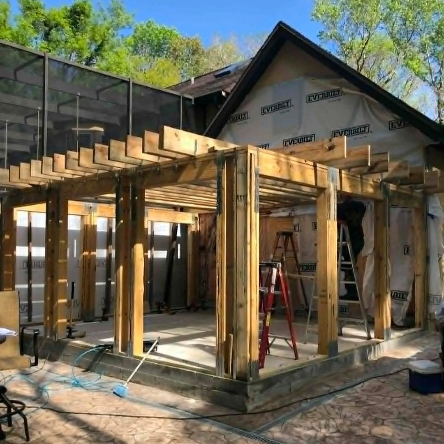 Construction site of a wooden frame house in progress, with wooden beams and ladders, surrounded by trees under a clear blue sky.
