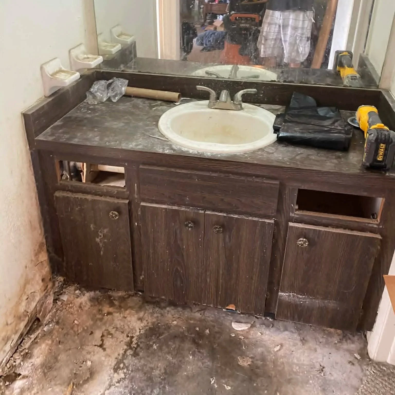 Dark wooden bathroom vanity with a white oval sink, two cabinet doors, and open storage space, covered in dust and debris, with tools on the countertop and a damaged wall behind it.