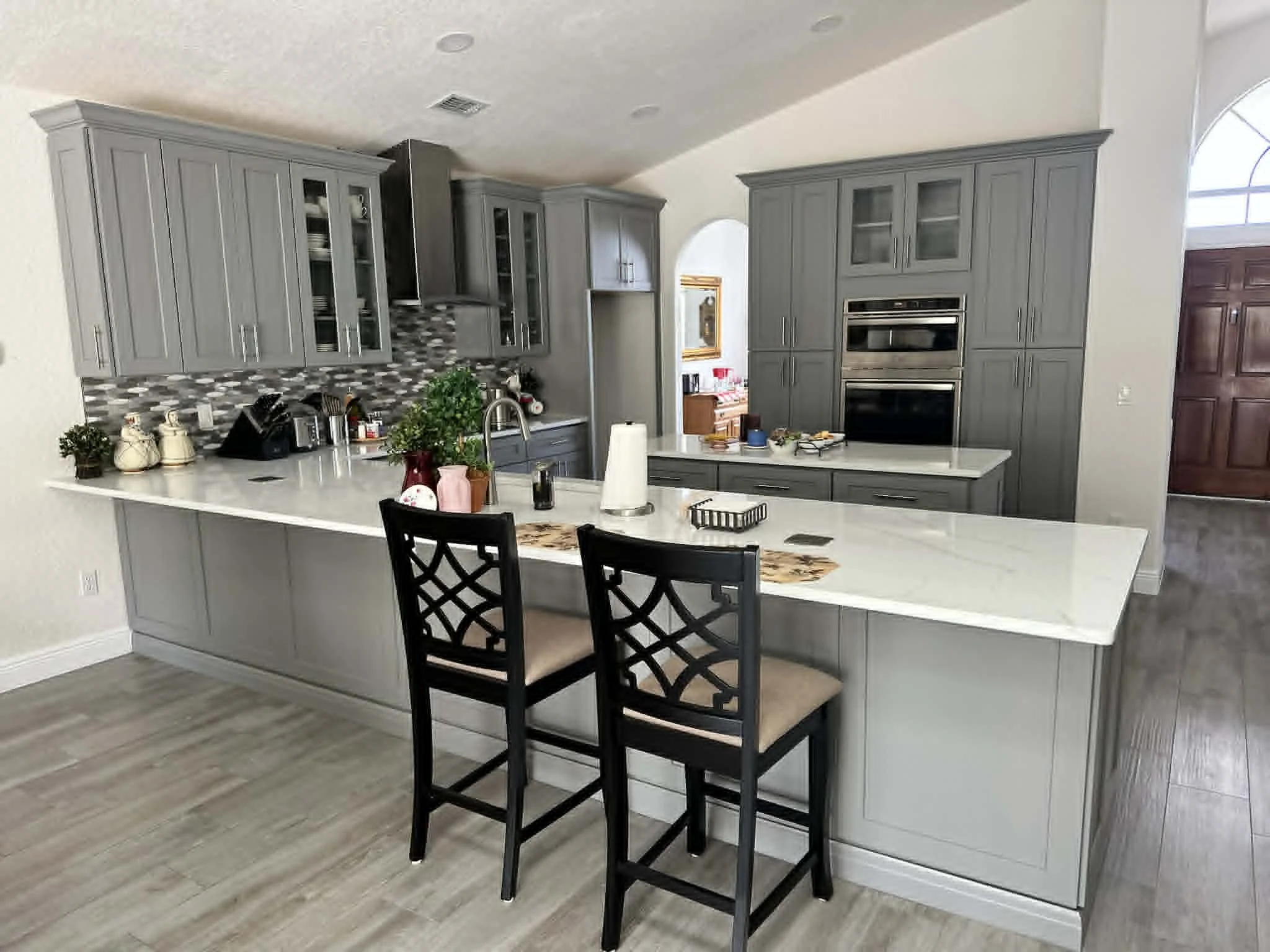 Modern kitchen with gray cabinets, a white countertop, and a mosaic tile backsplash. There are black chairs at the island, with various small items and plants on the counter.