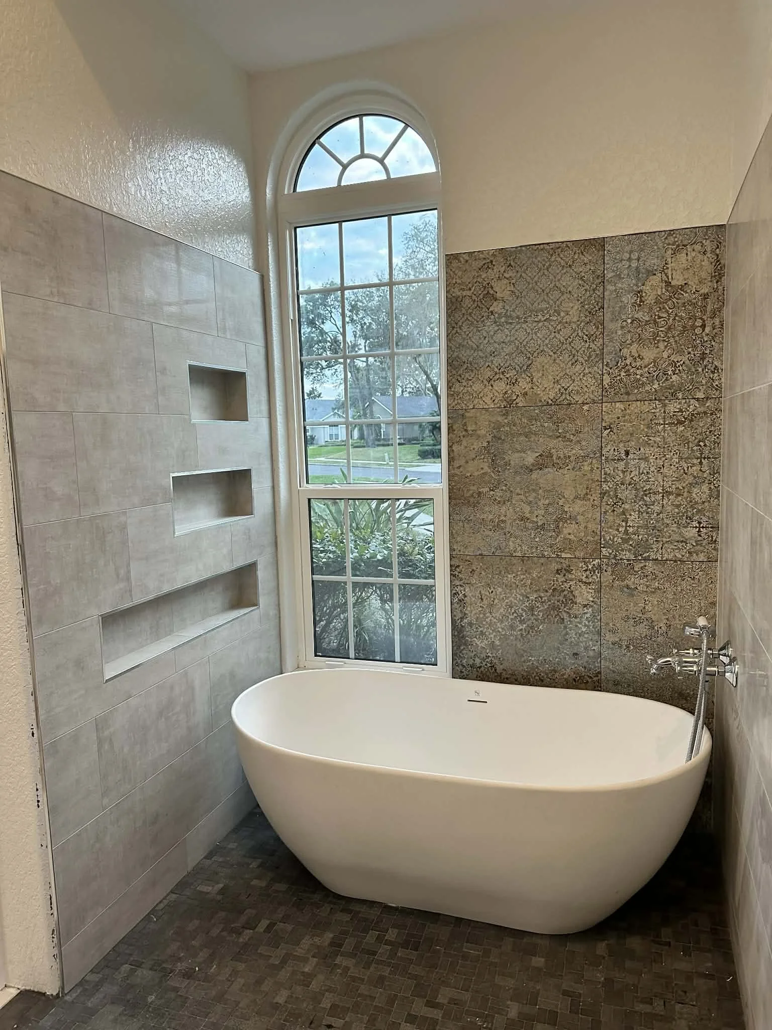 A modern bathroom with a large arched window, a white freestanding bathtub, tiled walls, and a view of a suburban street with trees outside.