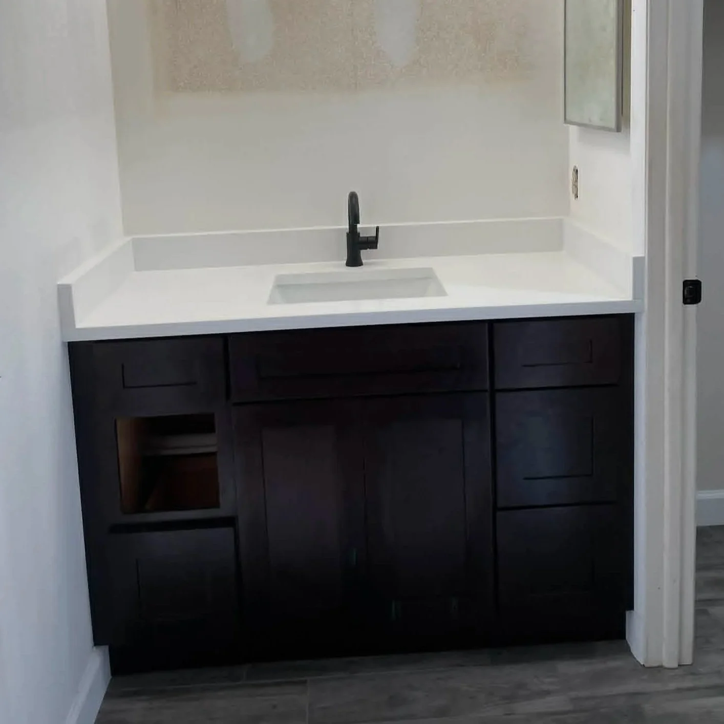 Bathroom vanity with white countertop, black faucet, and dark wood cabinet, mirror on wall, and gray flooring.