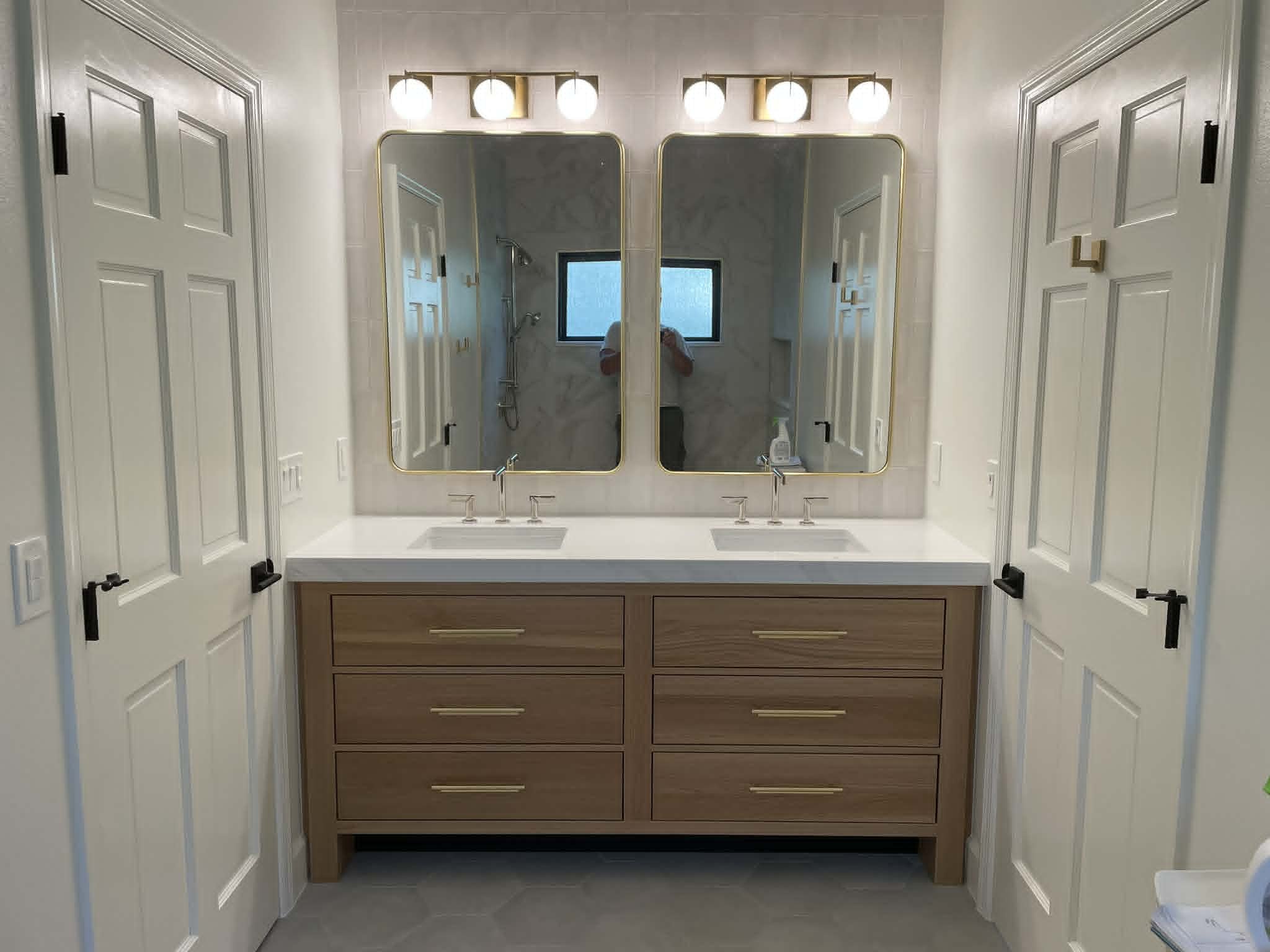 A bathroom with a double vanity with wooden drawers, large mirrors with gold trim, and three globe lights above each mirror. There are two white doors on either side, and a shower with a window can be seen in the reflection.