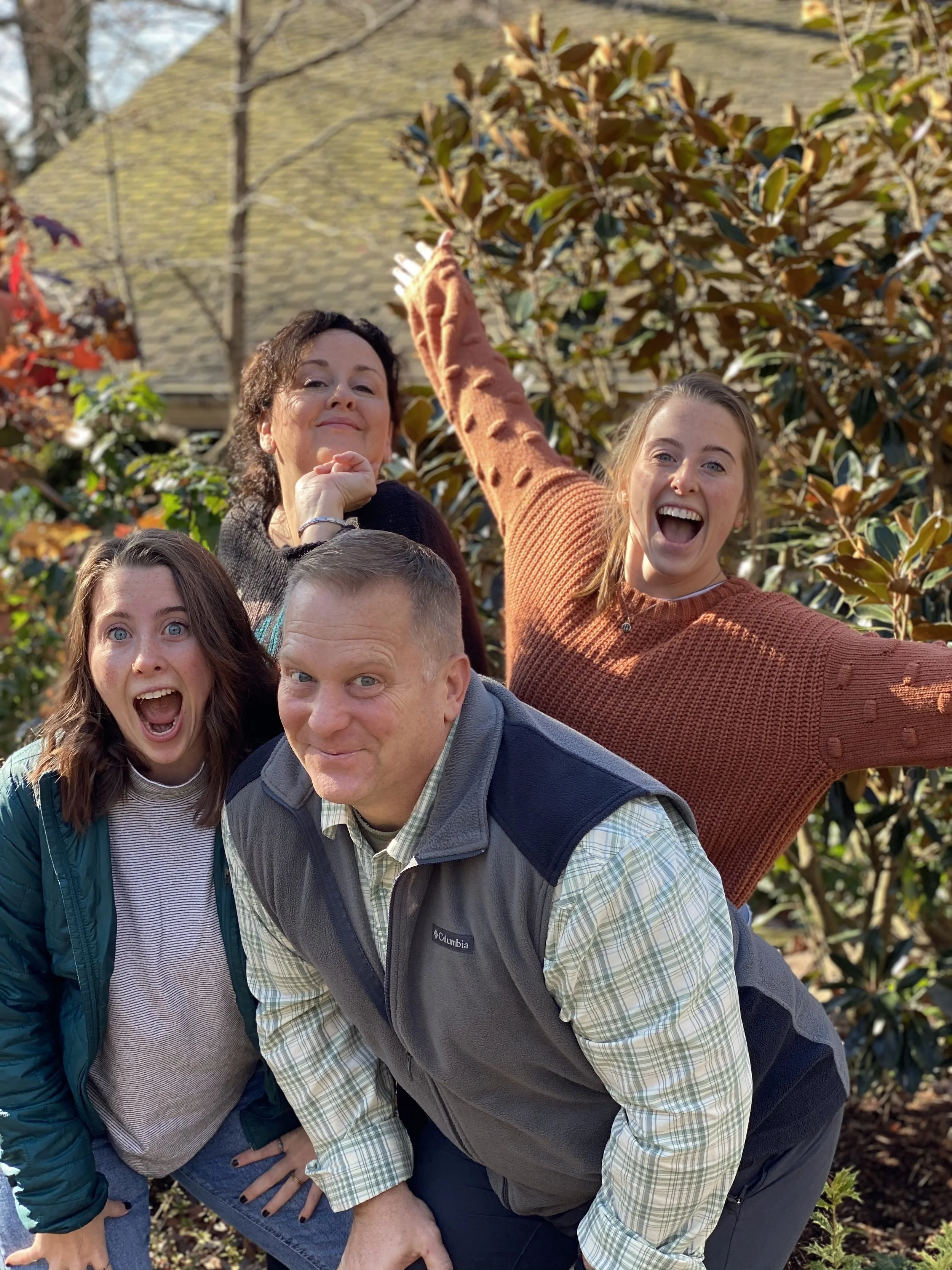 A group of five people outdoors, with two women in the back smiling and posing, one young woman in the front with open mouth, and a man and woman in the foreground smiling and leaning into each other, all surrounded by trees and plants.