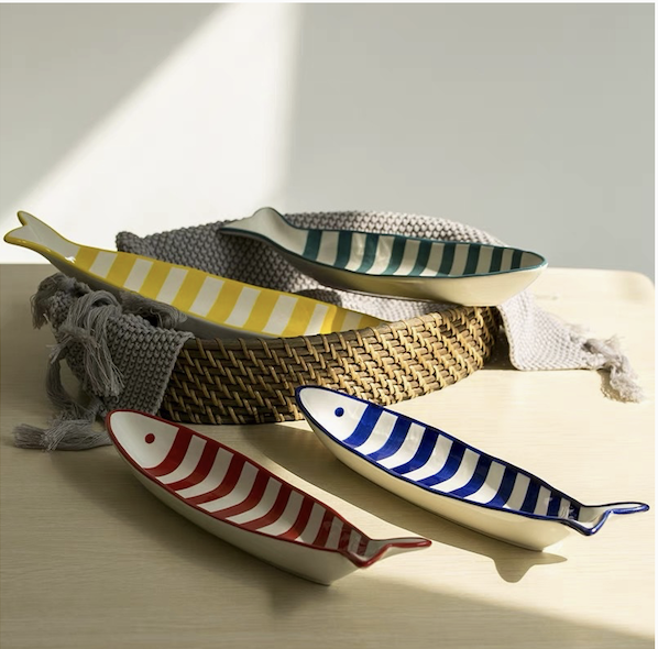 Four ceramic fish-shaped bowls with striped patterns in yellow, blue, red, and teal are arranged on a light wooden surface, with a woven basket and a gray textured cloth nearby.