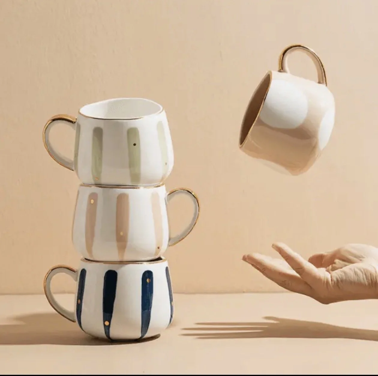 Three stacked white ceramic cups with gold rims and vertical stripes in gold, beige, and navy blue, with a hand balancing a similar cup in the air, against a plain beige background.