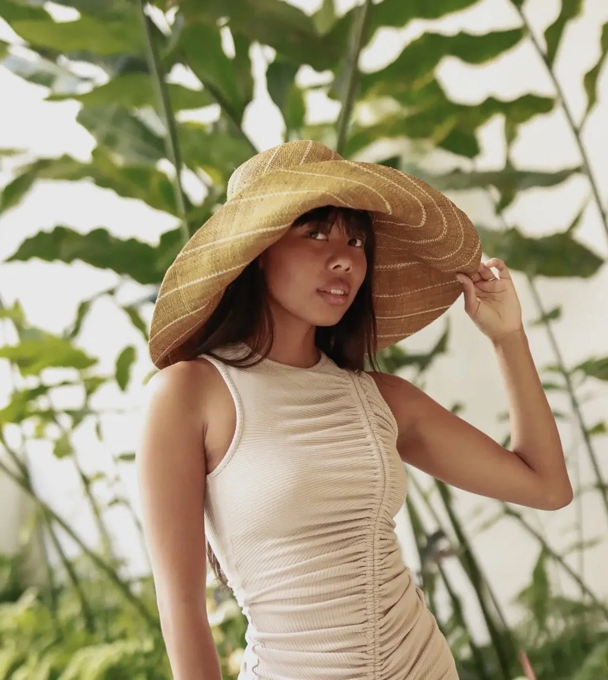 Young woman wearing a wide-brimmed straw hat , standing outdoors among green foliage.