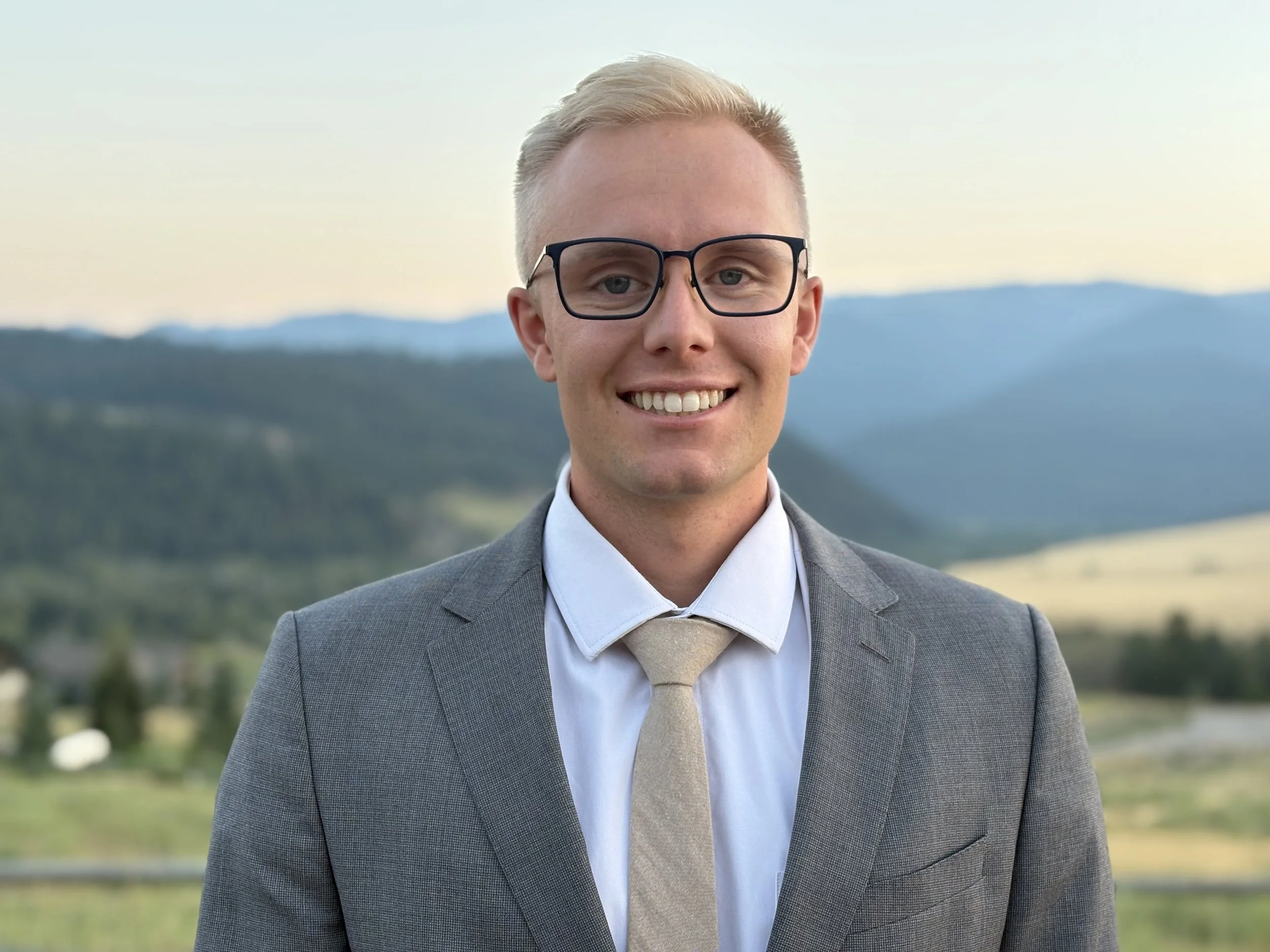 A young man in a gray suit with glasses, white shirt, and beige tie smiling outdoors with a mountain landscape in the background.
