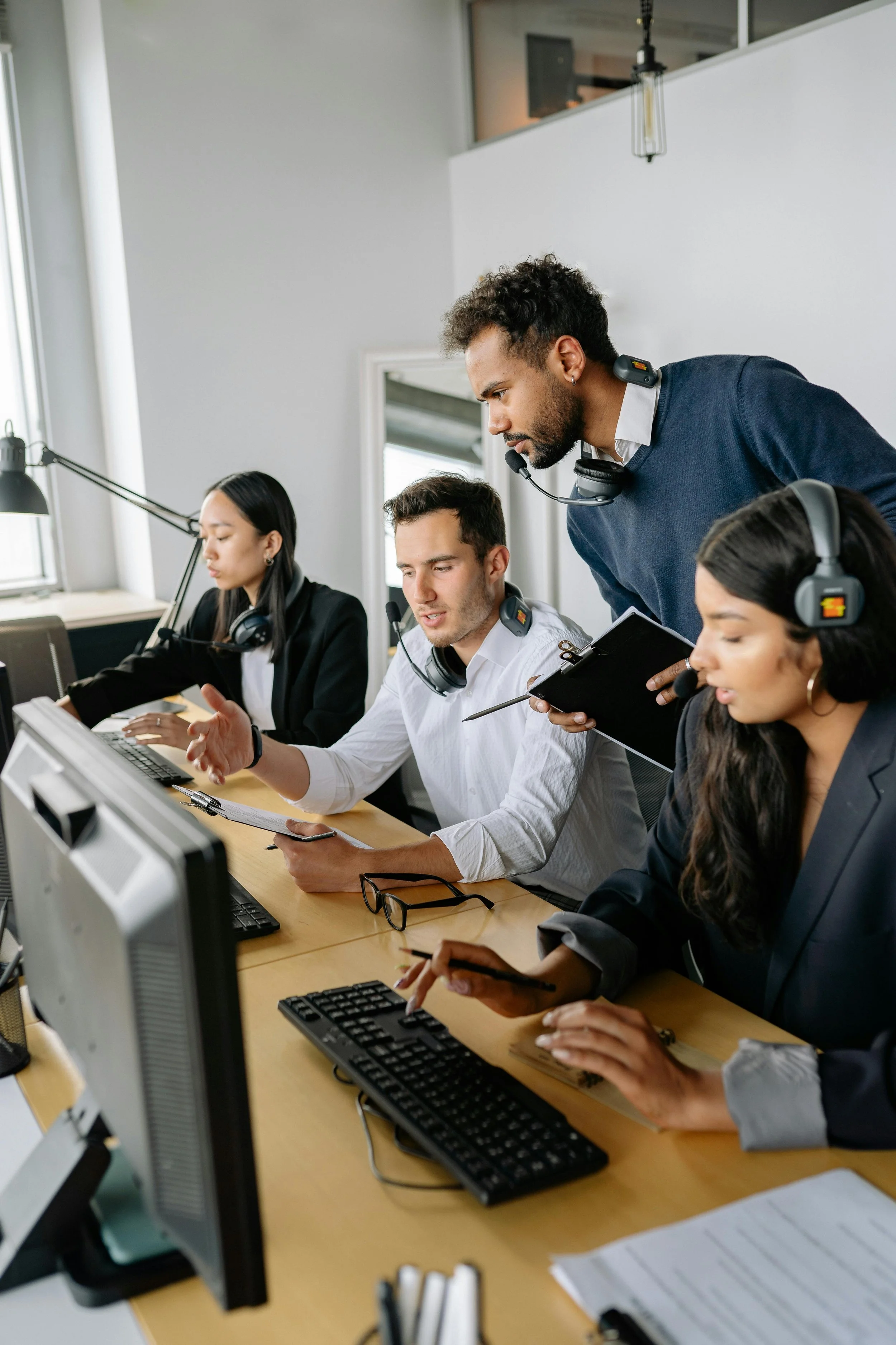 A diverse group of four customer service representatives working at their desks with headsets, computers, and paperwork in an office, engaged in a team discussion.