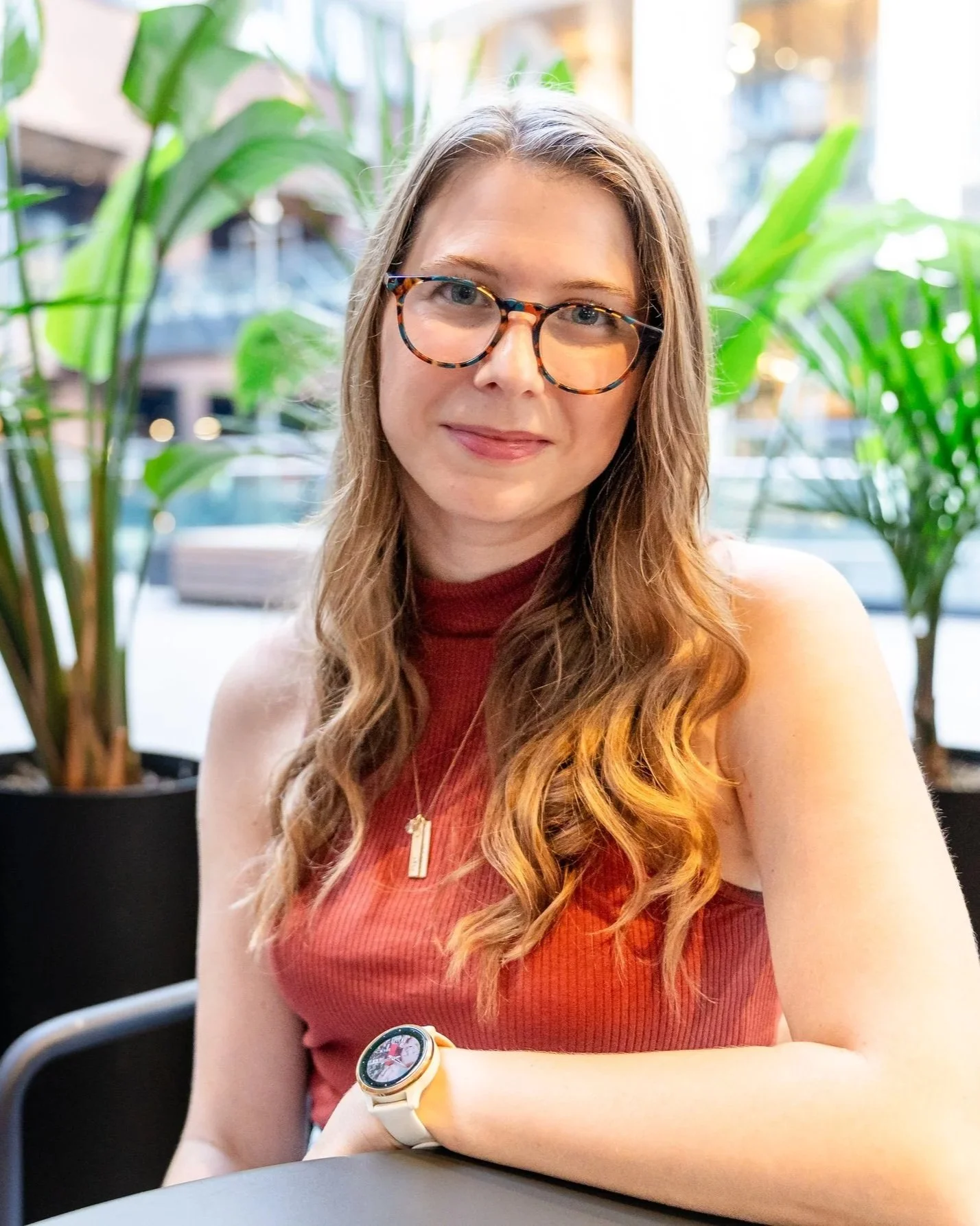 A woman with long wavy brown hair, wearing glasses, a red sleeveless top, a gold necklace, and a white smartwatch, seated at a table in a cafe with large green plants and a city street in the background.