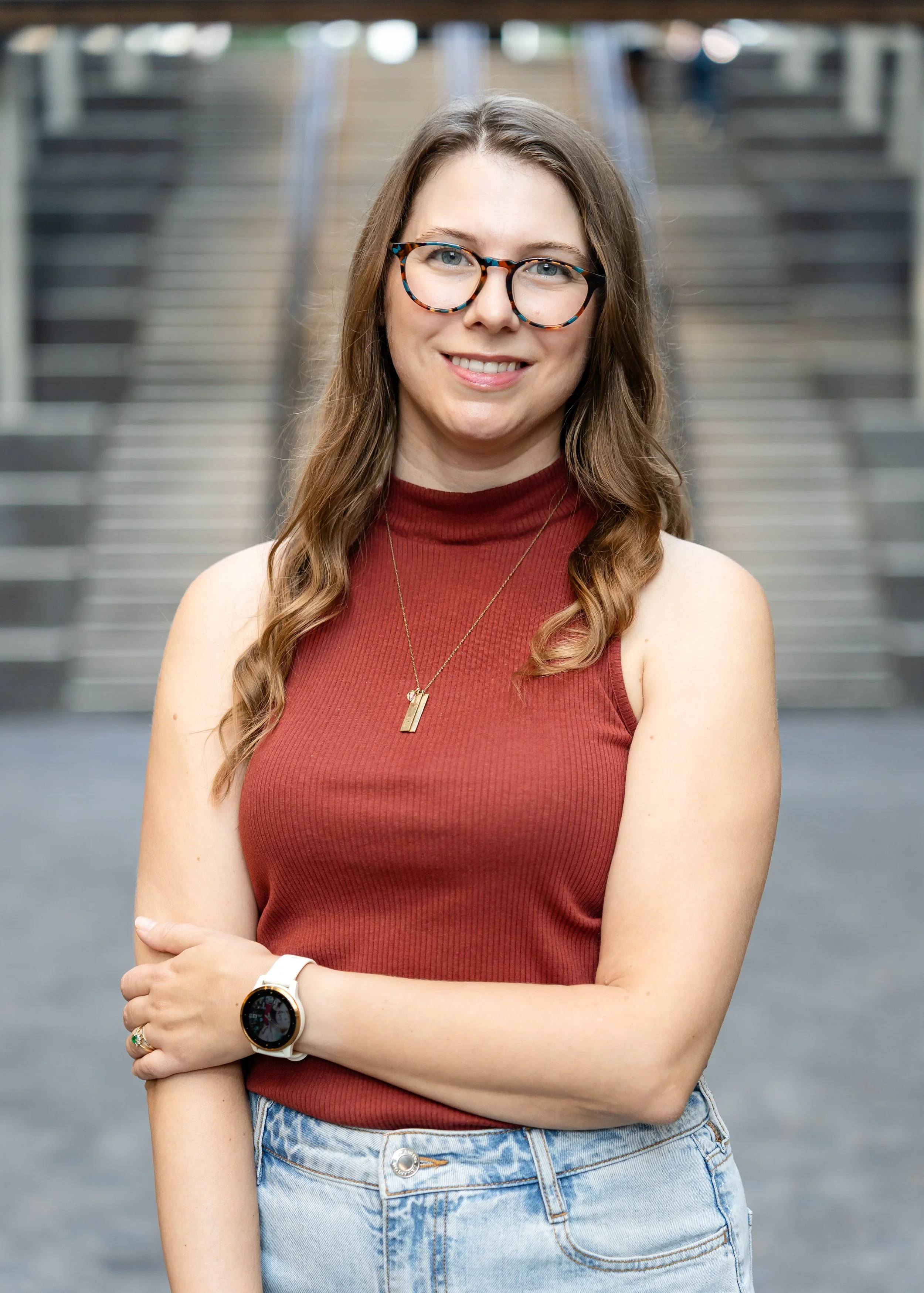 A woman with long wavy brown hair and glasses, smiling with arms crossed, standing in front of stairs at an indoor location.