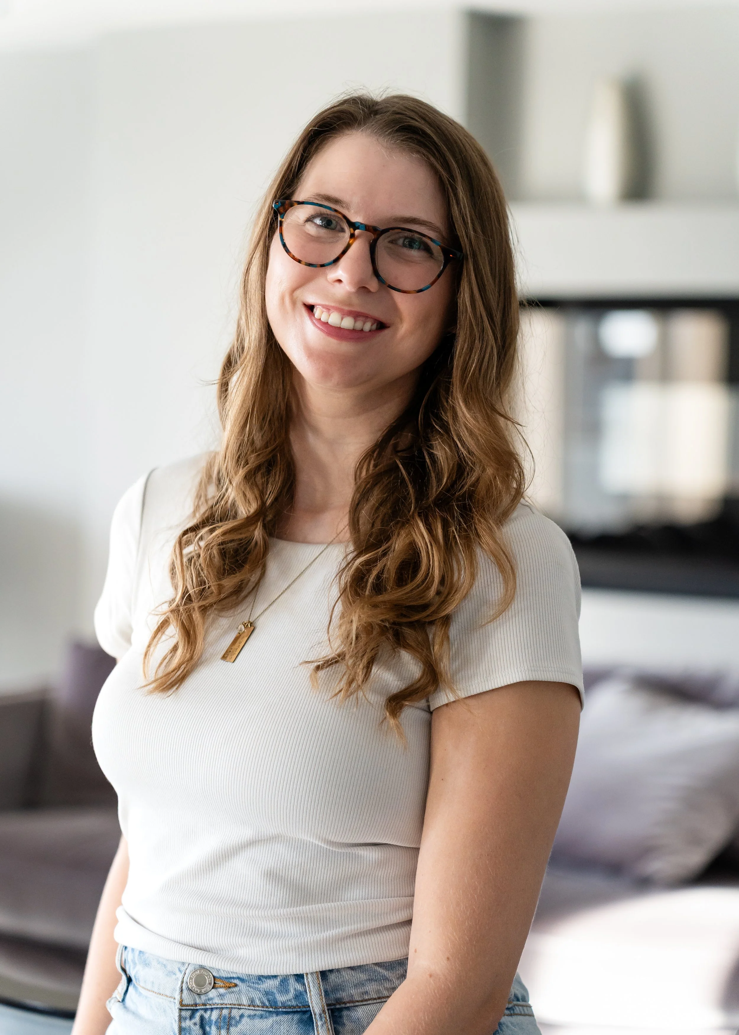 A woman with long, wavy brown hair, wearing glasses, a white shirt, and a necklace, smiling indoors with a blurred background.