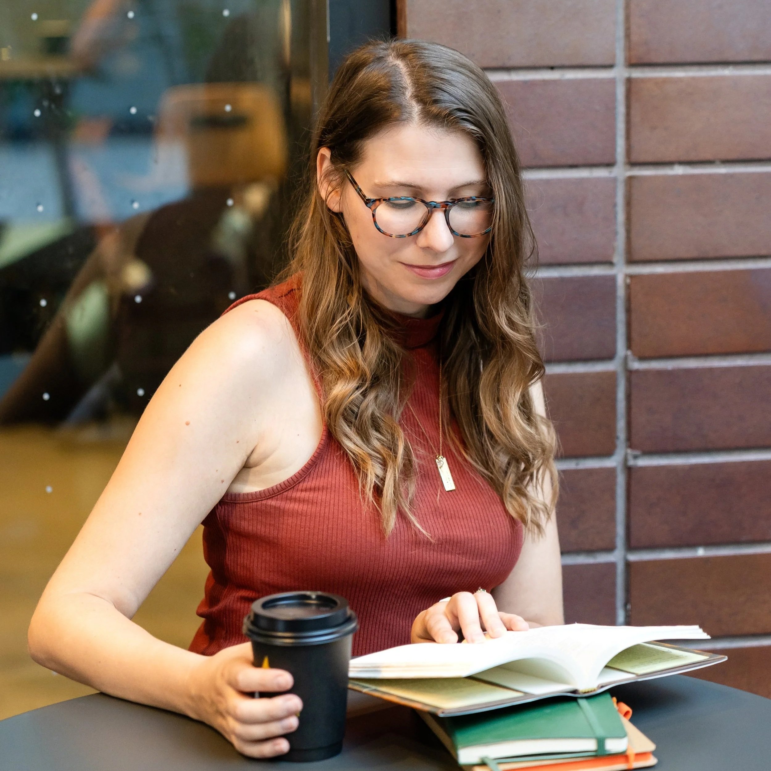A woman with glasses sitting at a table, reading a book, holding a coffee cup, with a brick wall behind her.