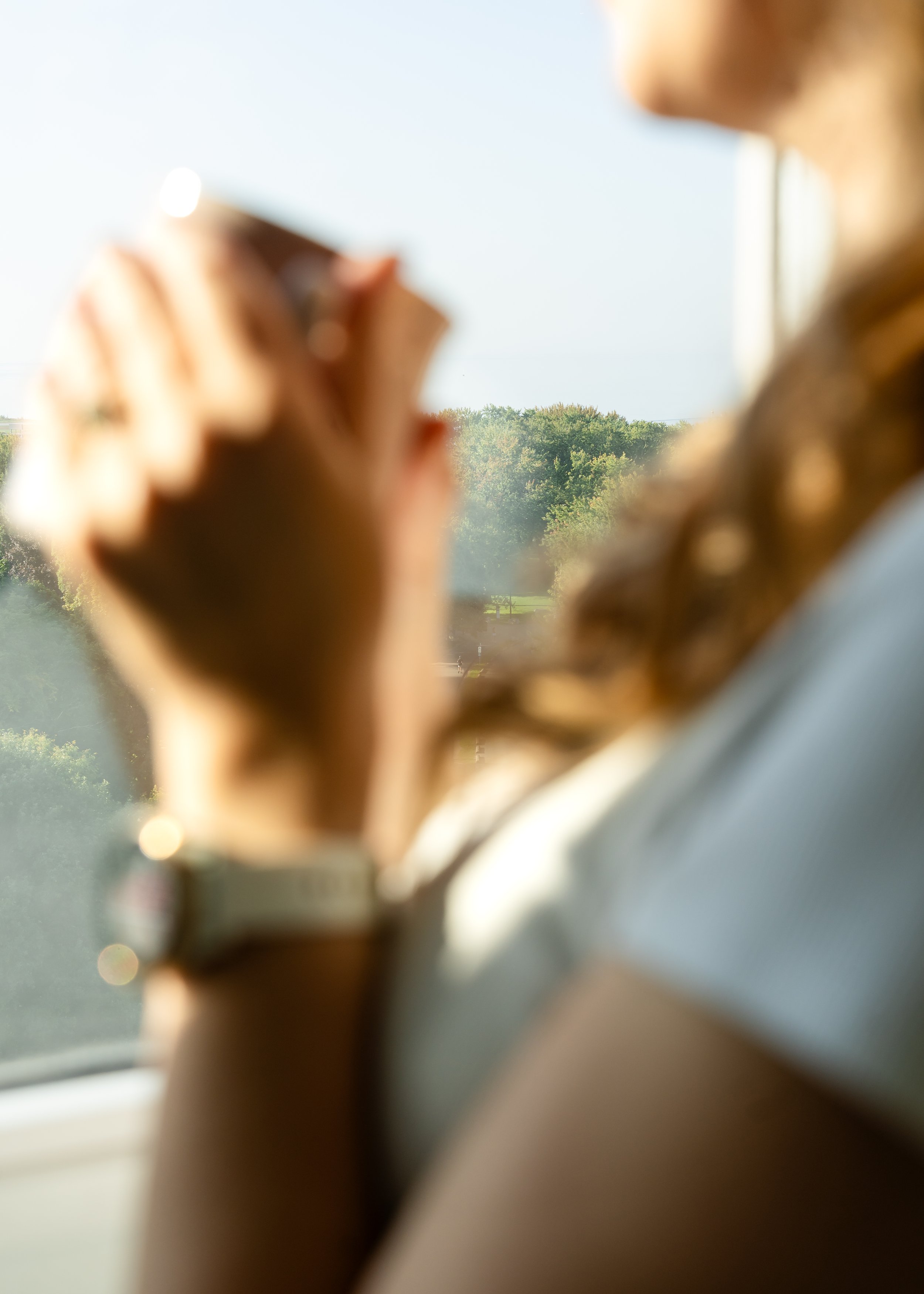 Person holding a cup of coffee with a park and trees in the background, seen through a window.