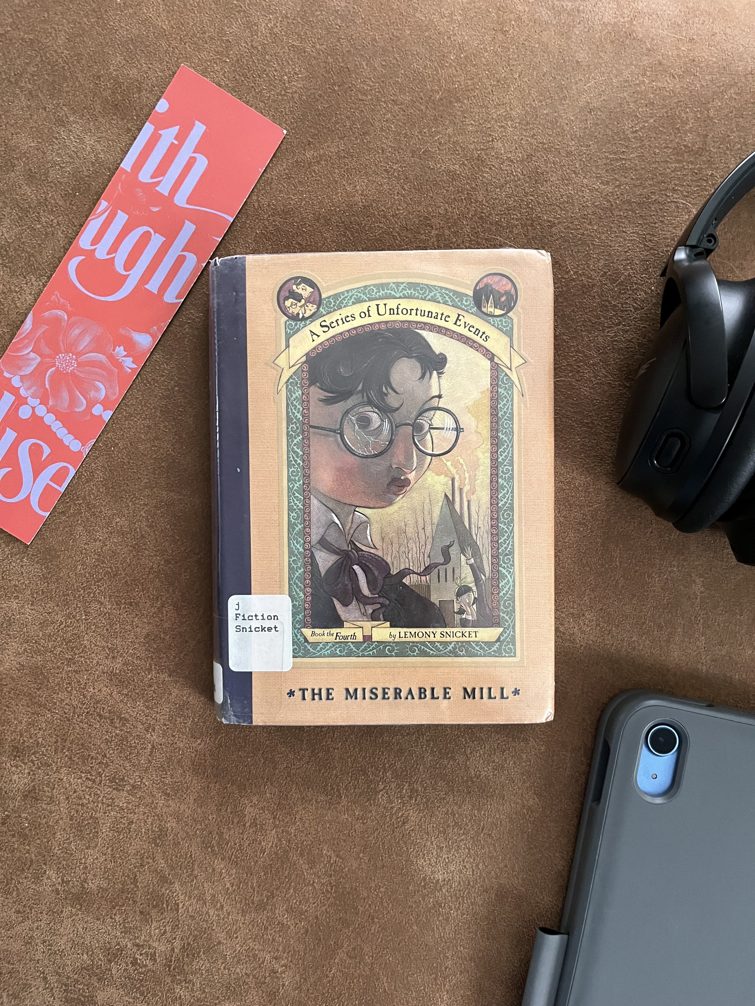 A book titled 'The Miserable Mill' by Lemony Snicket from the 'Series of Unfortunate Events' stands on a brown surface. To the right is a pair of black headphones and a gray phone, and to the left is a red and blue bookmark.